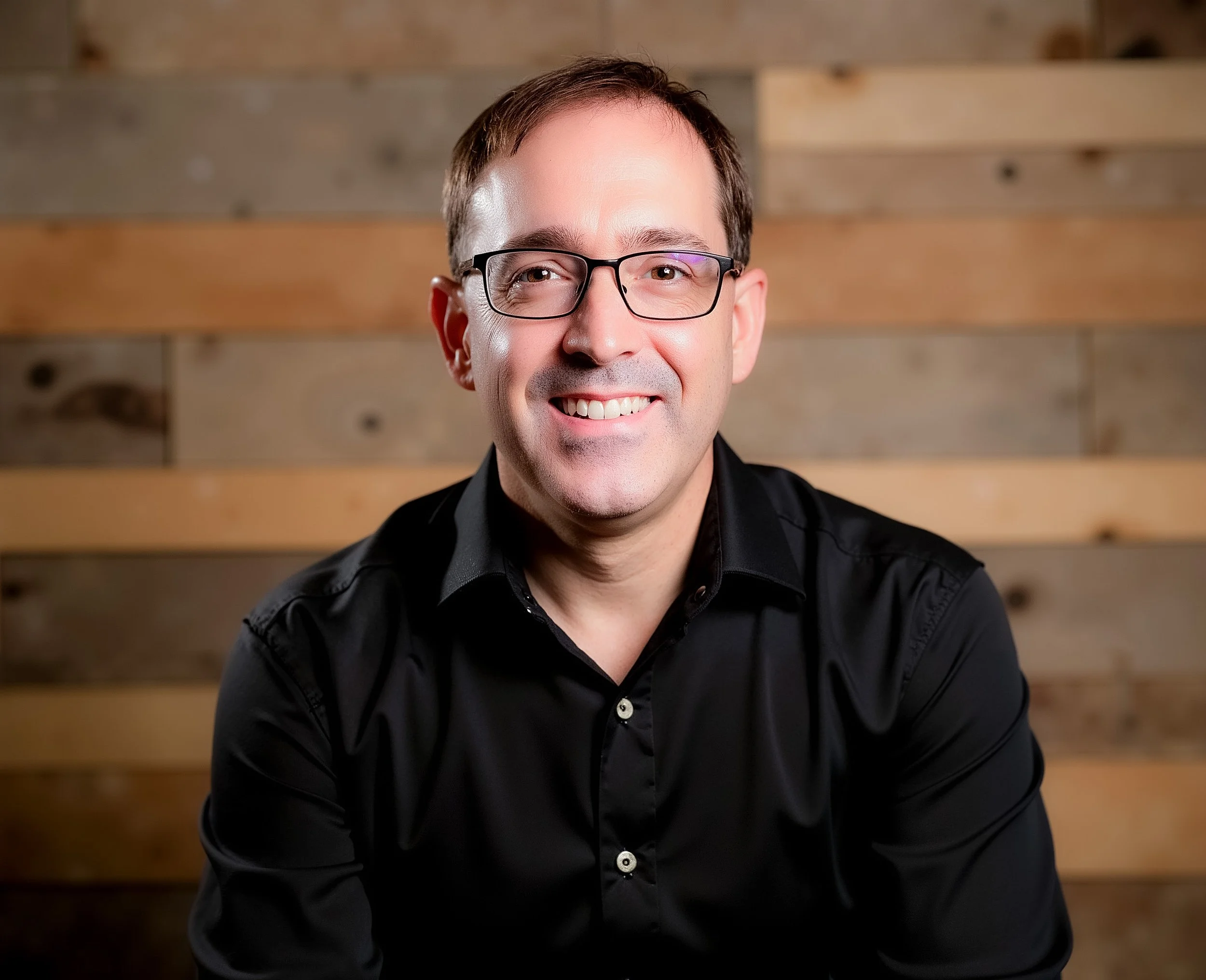 A smiling man with glasses wearing a black shirt in front of a wooden background.
