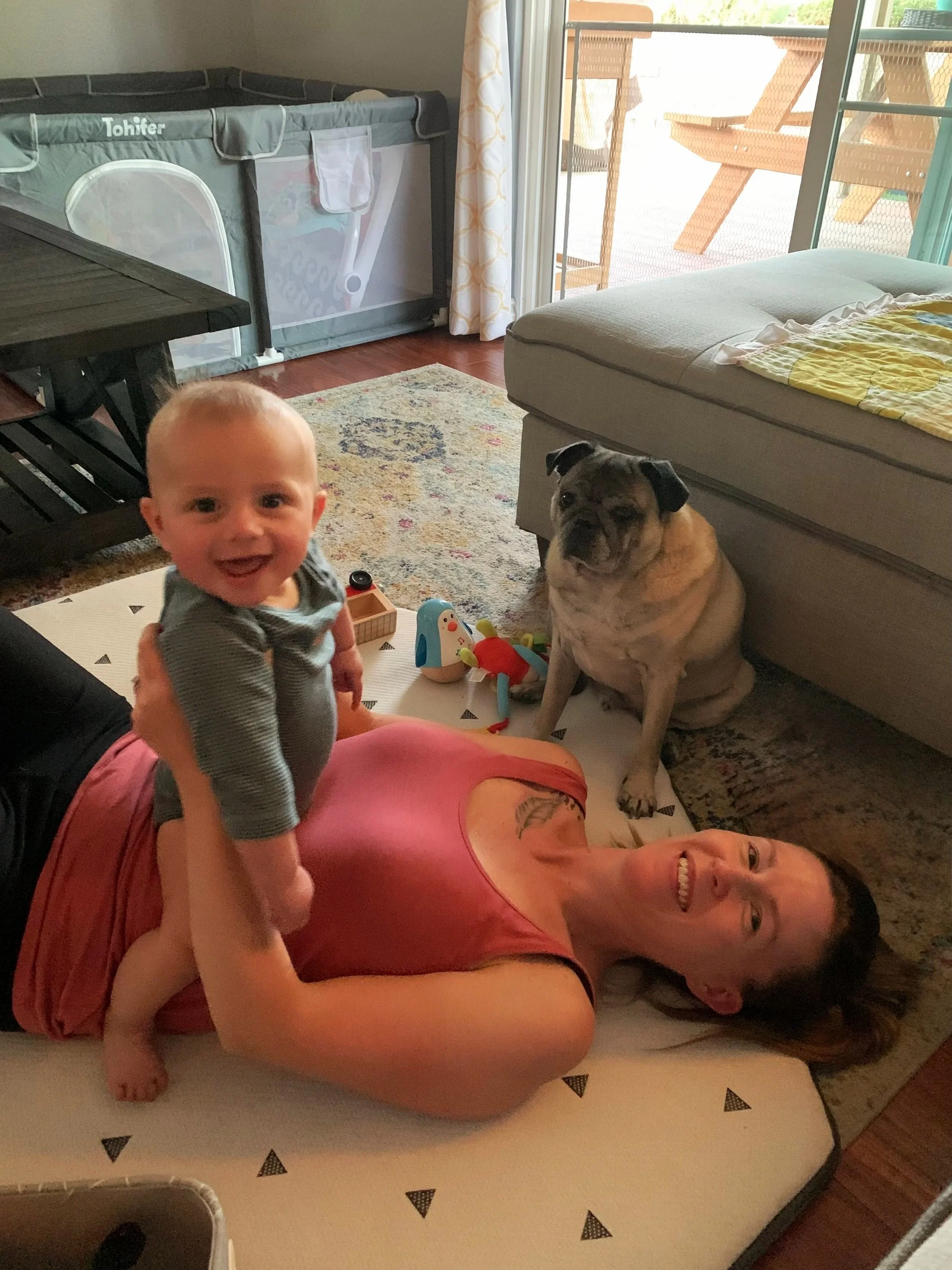 A woman lying on a bed, smiling, with a baby standing on her stomach, and a bulldog sitting nearby in a room with a sliding glass door leading to a balcony.