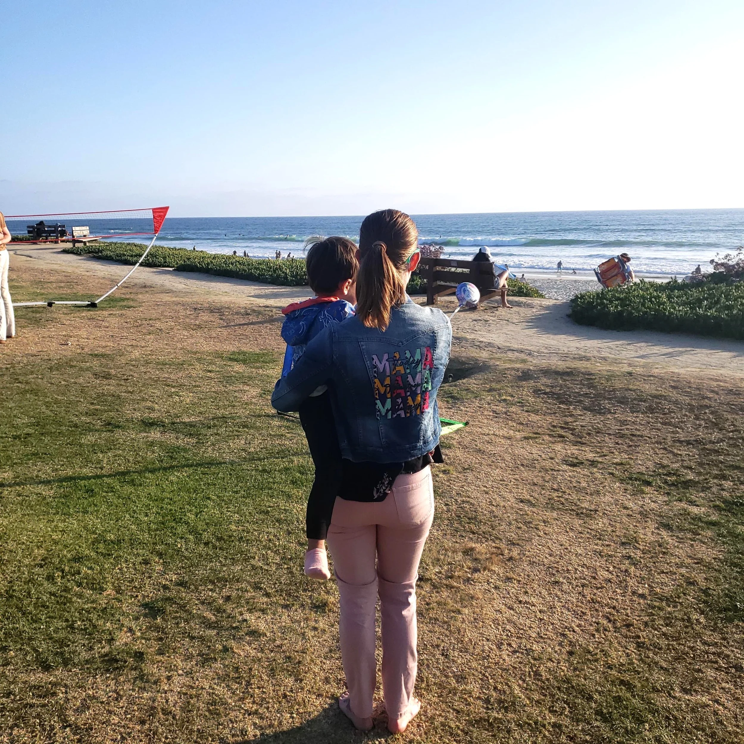 A woman and a young girl walking on a grassy beach area near the ocean, with benches and a lifeguard tower visible in the background. They are dressed casually, and the scene is lit by sunlight, suggesting late afternoon or early evening.
