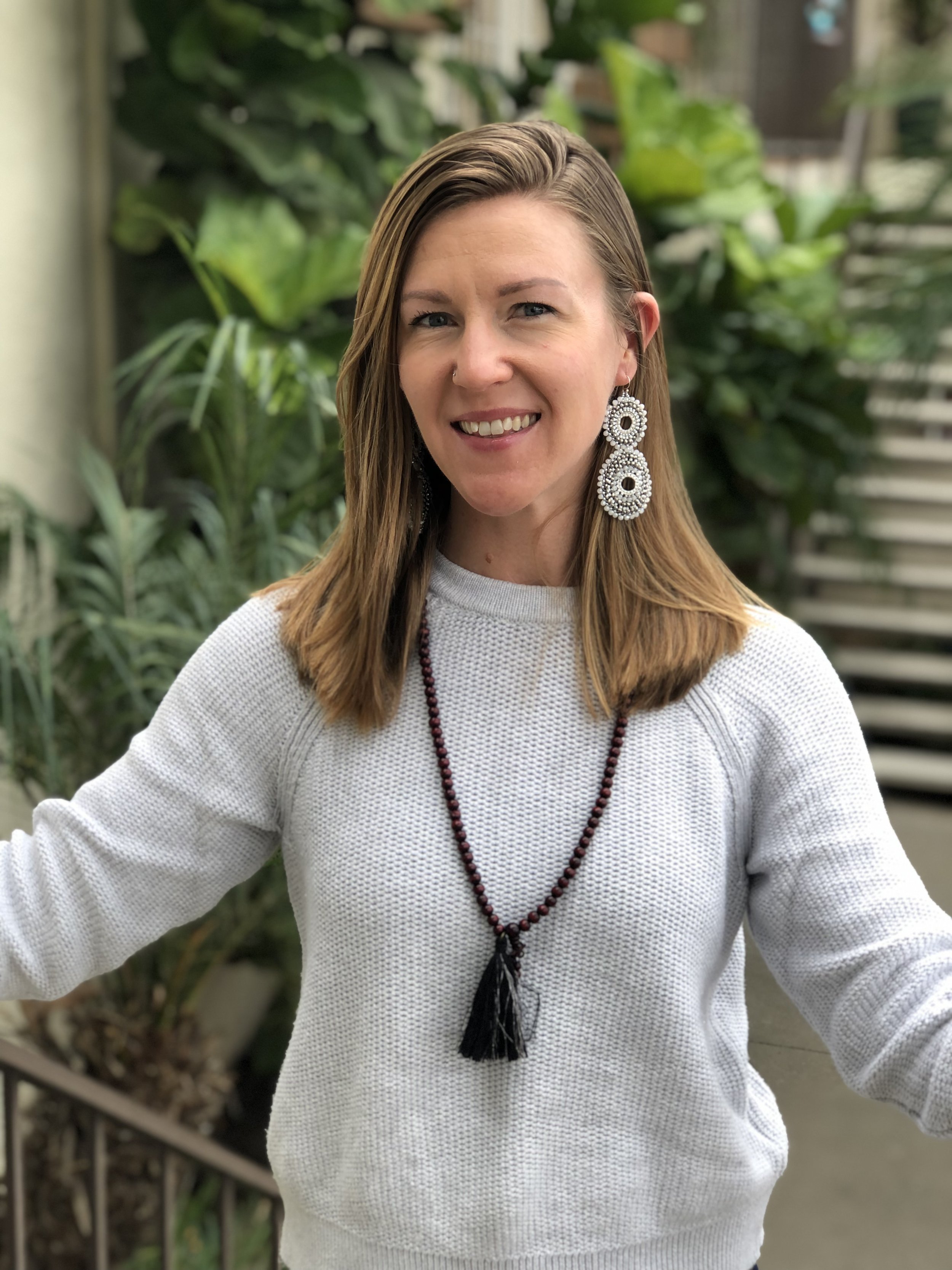 A woman with light brown hair wearing a white sweater, large white beaded earrings, and a beaded necklace with a tassel, smiling with greenery behind her.