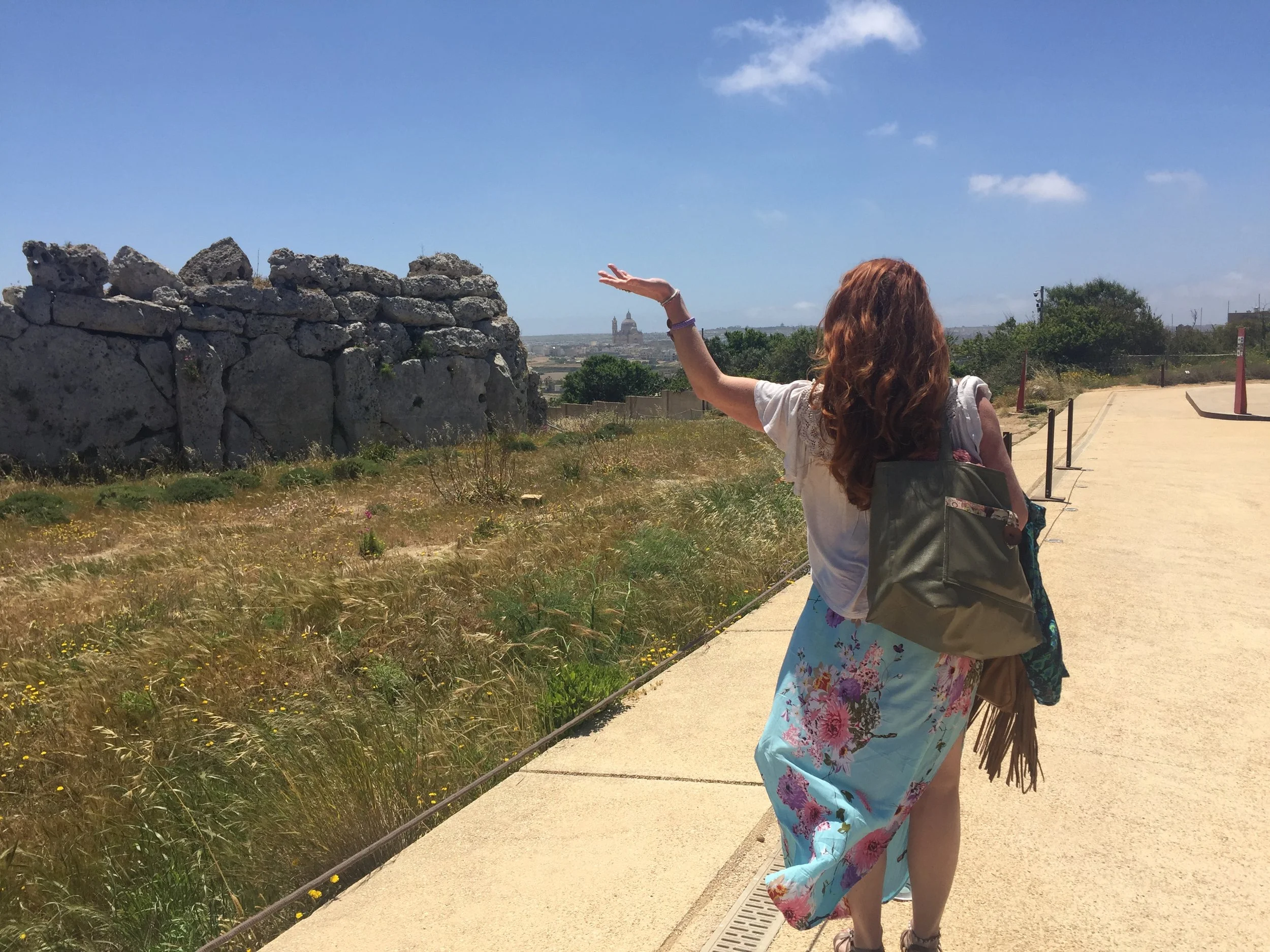 A woman with long red hair wearing a floral skirt and a white blouse holding a beach bag and walking along a sandy path with her arm outstretched, near a stone wall and trees, with a historic building in the distance under a partly cloudy sky.
