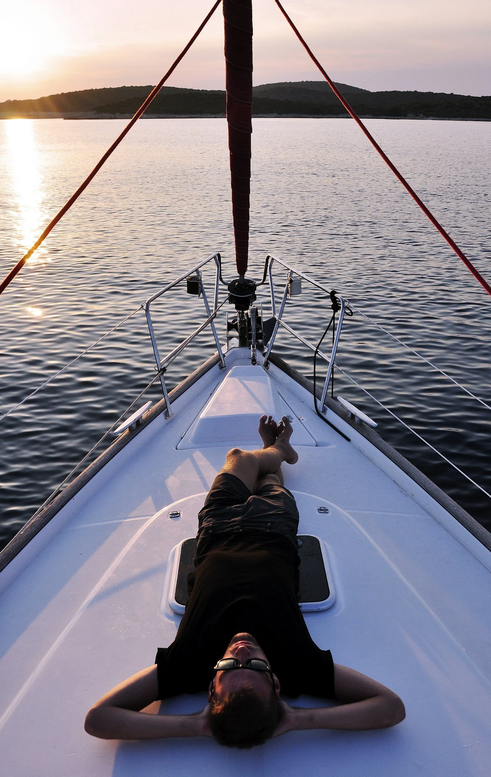Person lying on the deck of a boat during sunset, with a view of the water and distant land in the background.