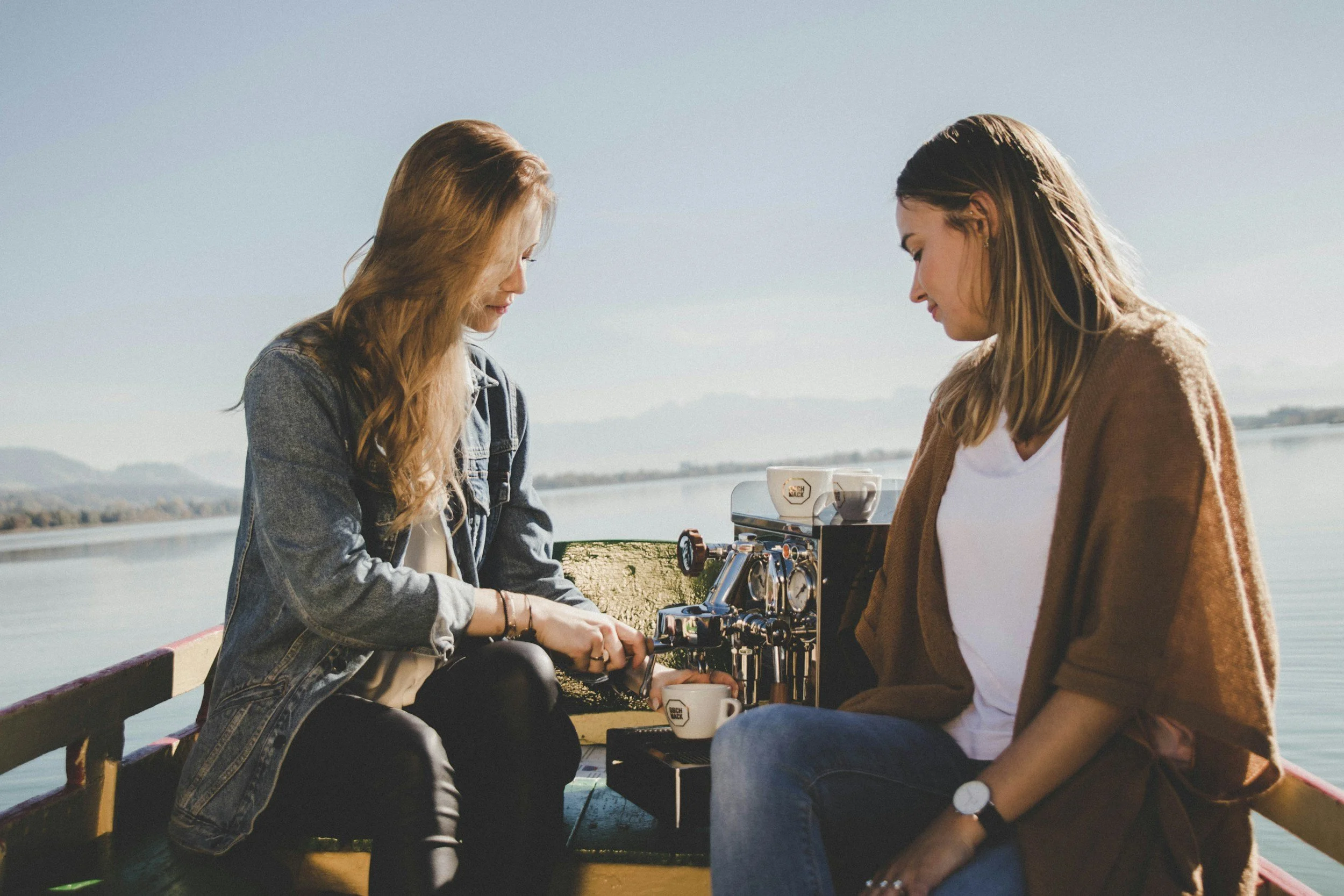 Two young women sitting on a dock by a body of water, preparing to make coffee with a portable espresso machine. The sky is clear and the scenery includes distant mountains.