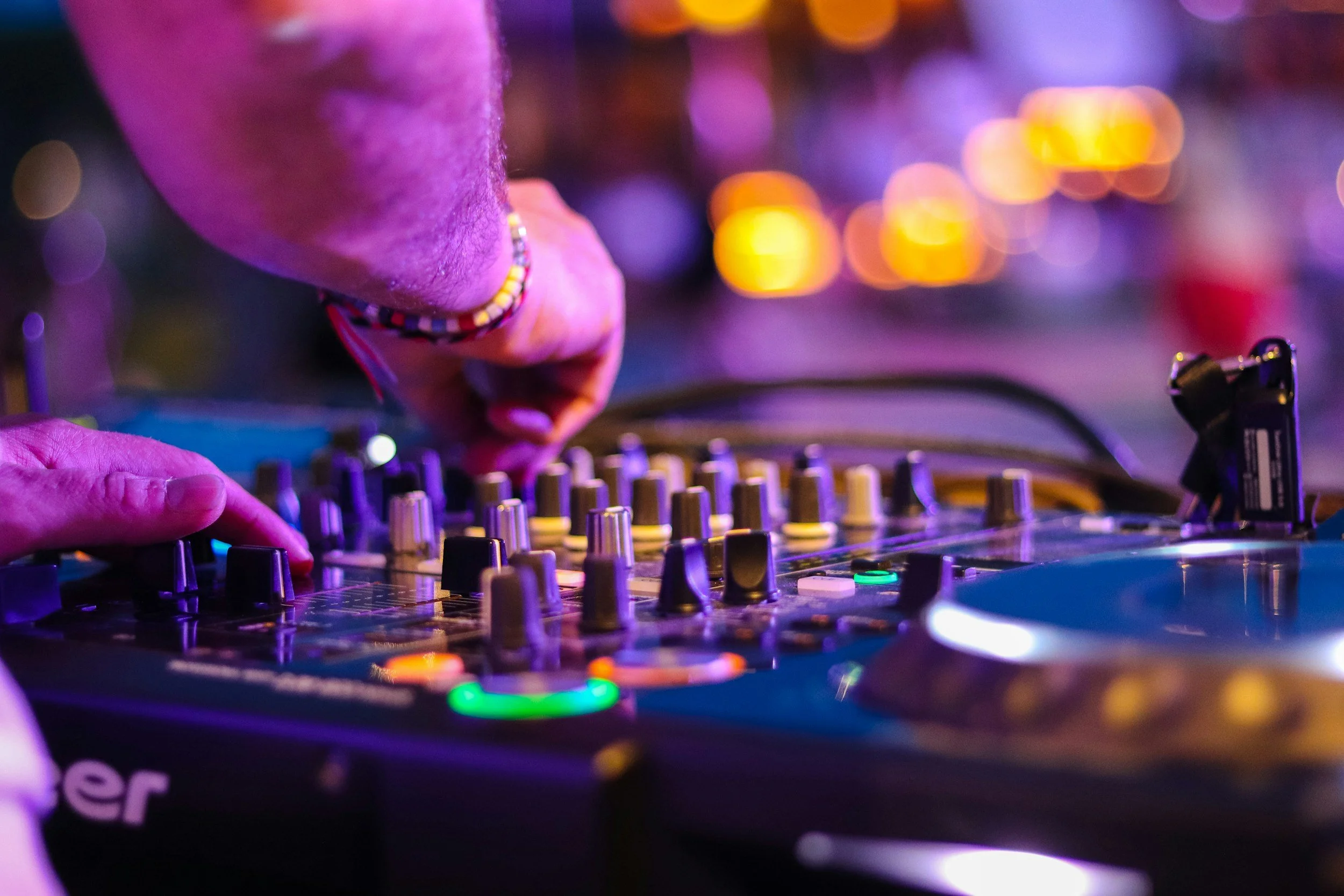 Close-up of a DJ's hand adjusting the knobs on a sound mixing console, with colorful, blurred lights in the background.