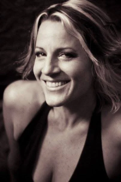 A woman with short, wavy blonde hair smiling and winking, wearing a sleeveless top.