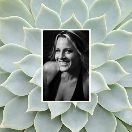 Black and white portrait of a smiling woman with shoulder-length hair, set against a background of green succulent plant leaves.