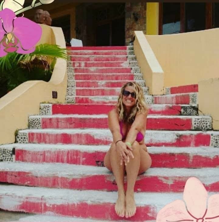 Woman sitting on colorful steps outdoors, wearing sunglasses and a purple top, smiling.