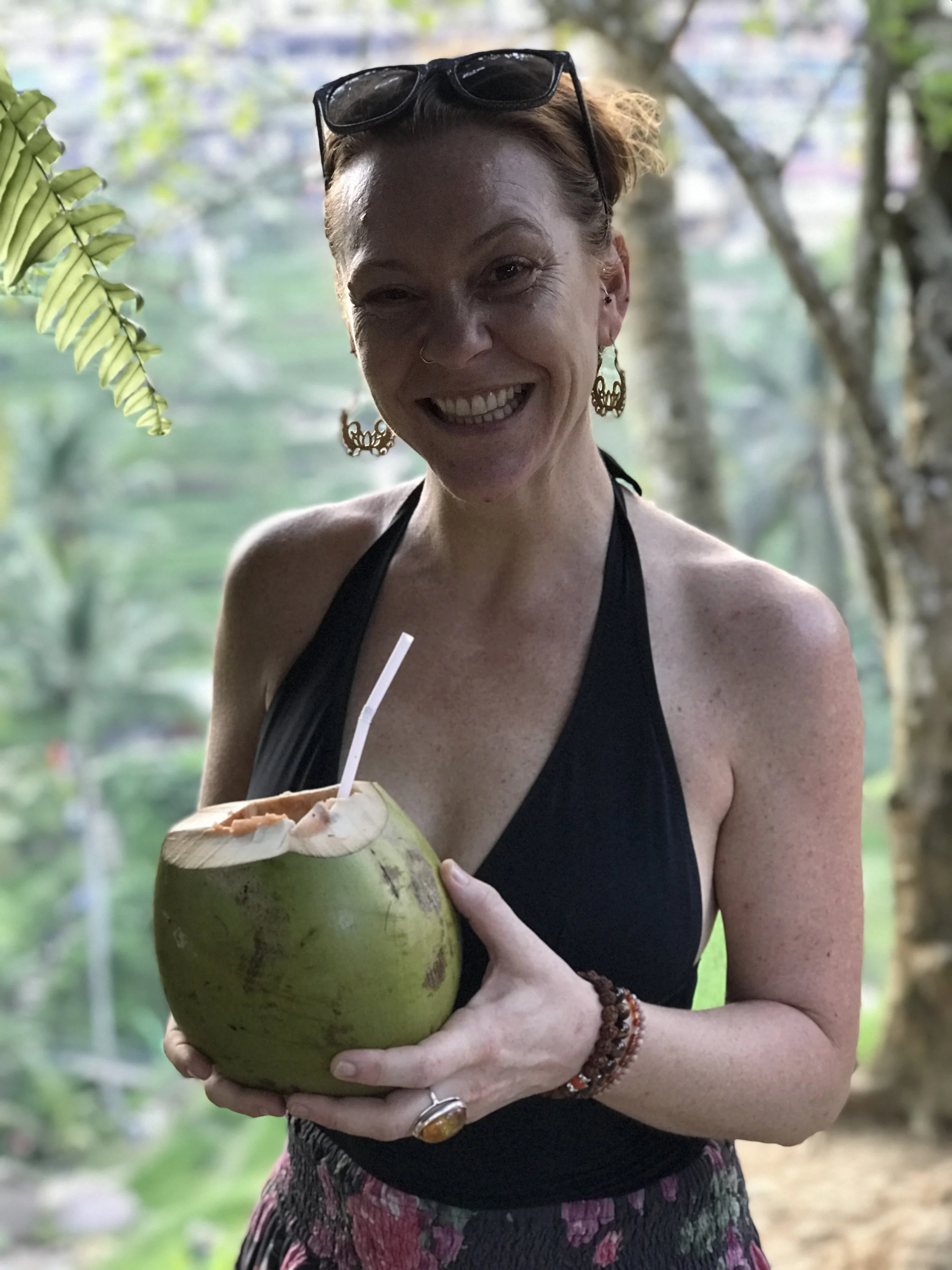 A woman with short hair, wearing glasses on her head, earrings, and jewelry, smiling while holding a green coconut with a straw inside, outdoors with trees in the background.