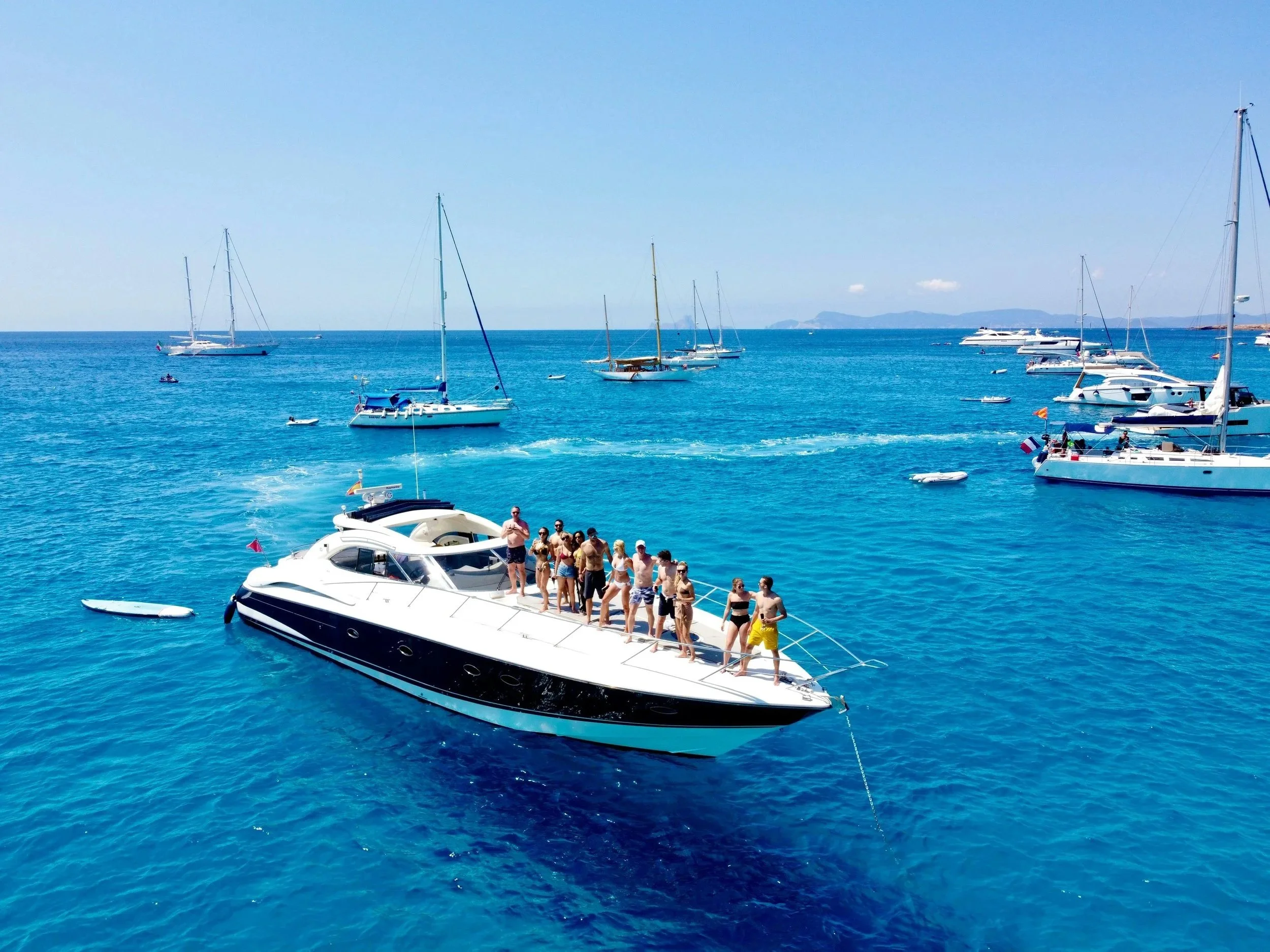 People on a large yacht floating in clear blue water with several sailboats and yachts anchored nearby, and a distant landmass on the horizon.