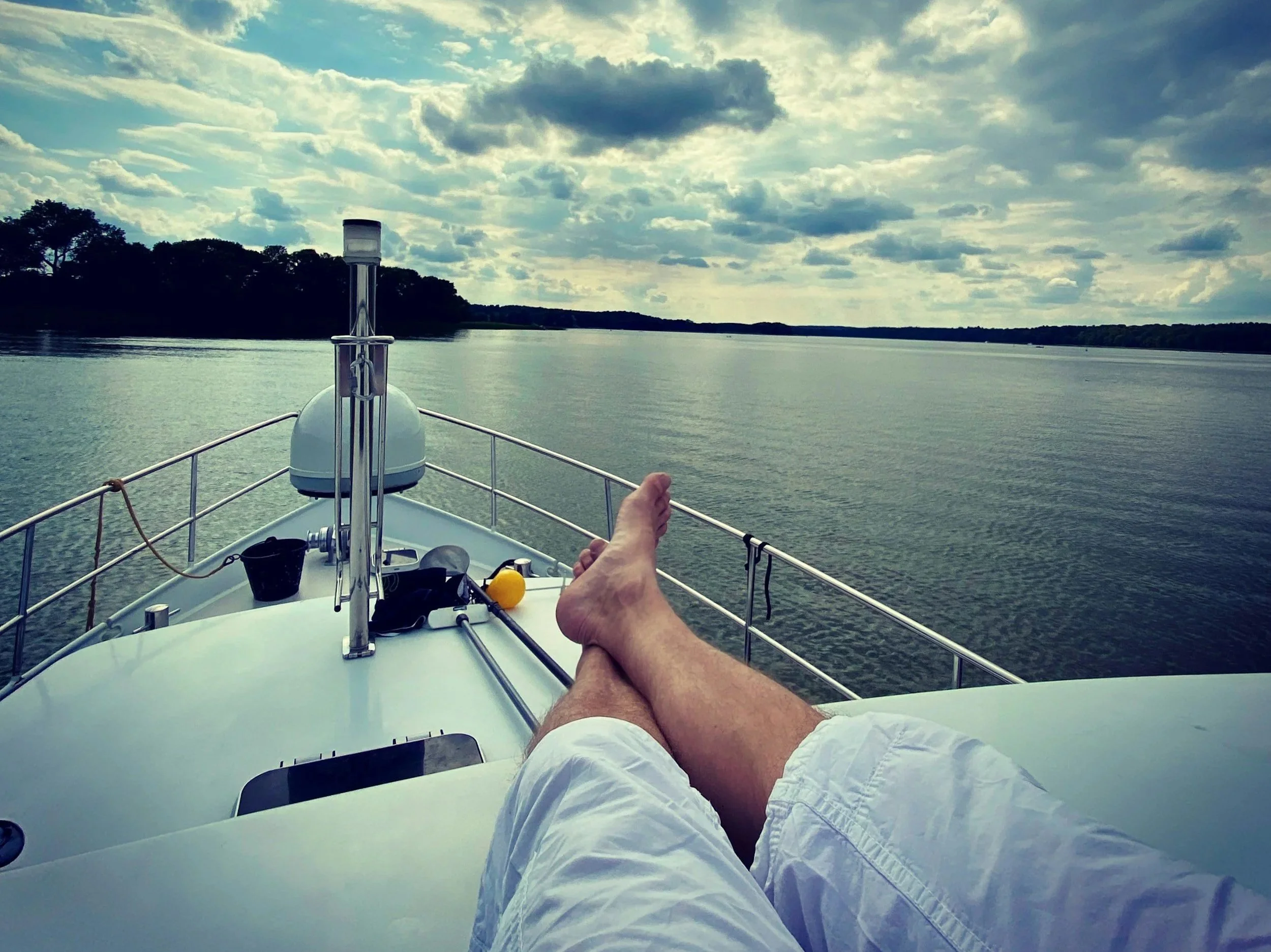 A person relaxing on a boat with their legs outstretched, view of water and cloudy sky, silhouette of trees in the distance.