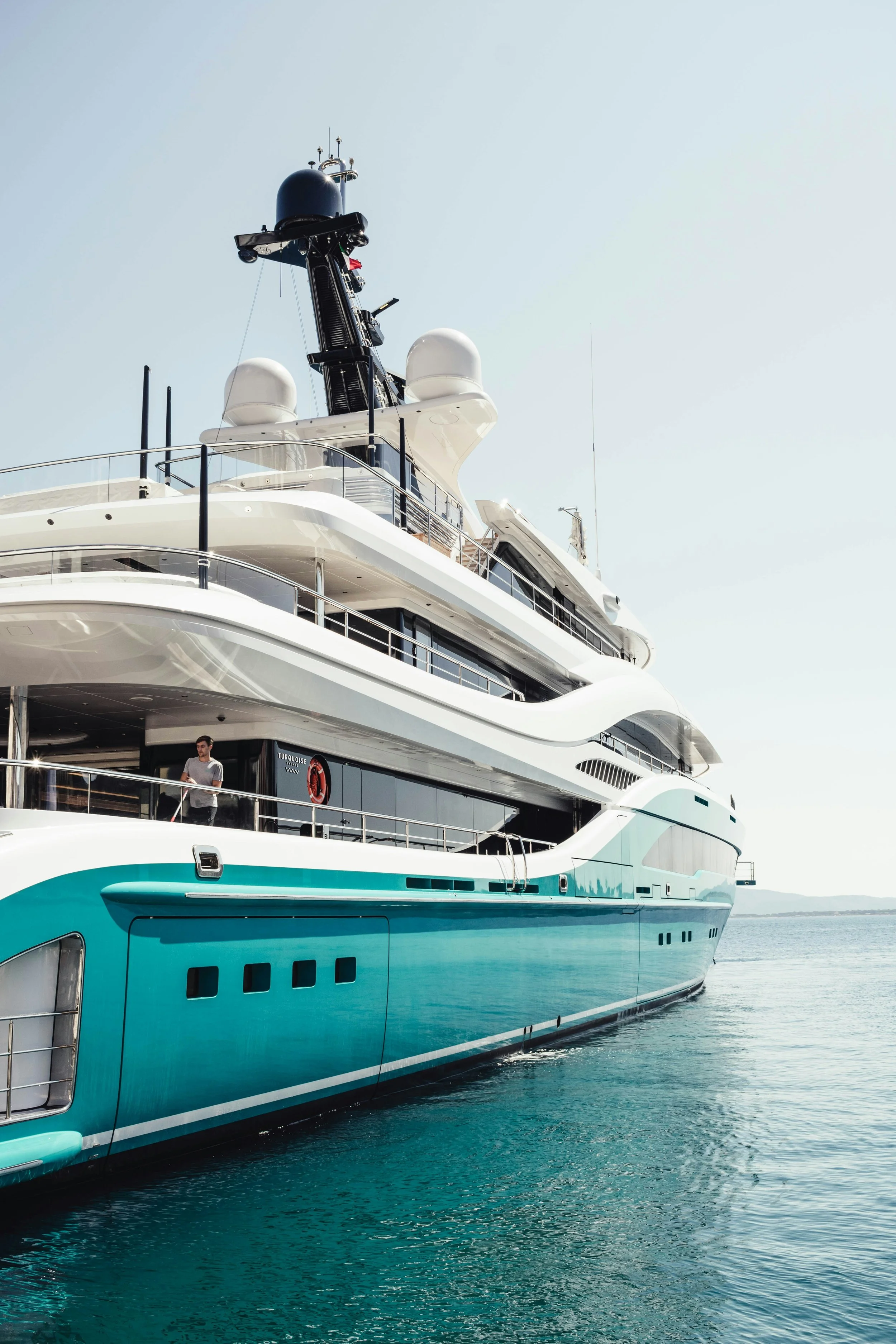 A large luxury yacht with a white and turquoise exterior docked on the water, showing multiple decks and a man standing on one of the lower decks.