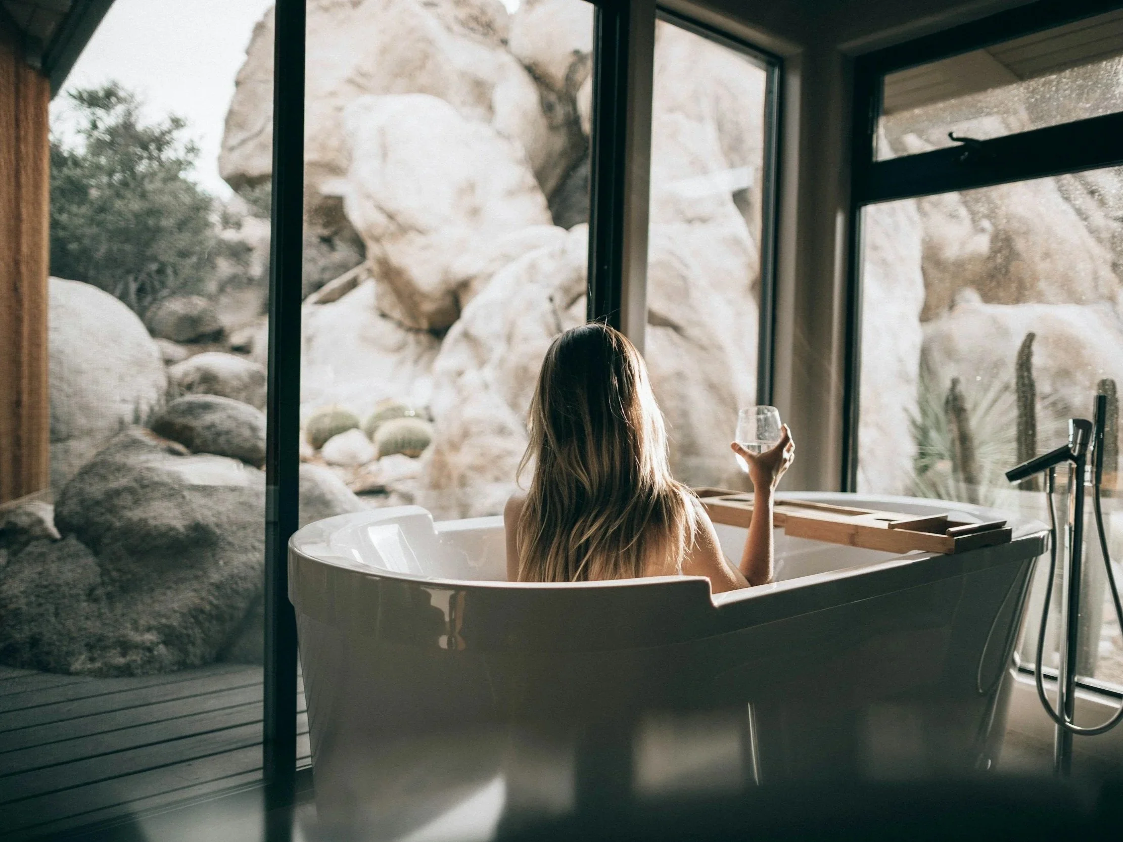 A person with long hair sitting in a bathtub holding a glass of water, with rocky outdoor scenery visible through large windows nearby.