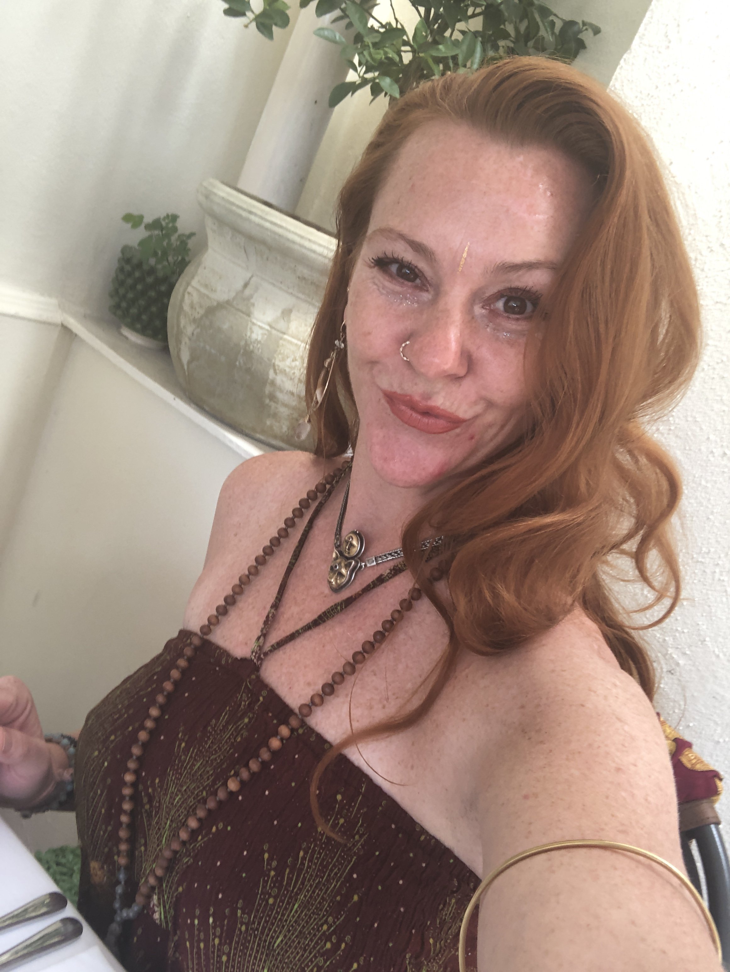 A red-haired woman with piercings, wearing necklaces and jewelry, takes a selfie at a decorated table with a plant and a large ceramic vase in the background.