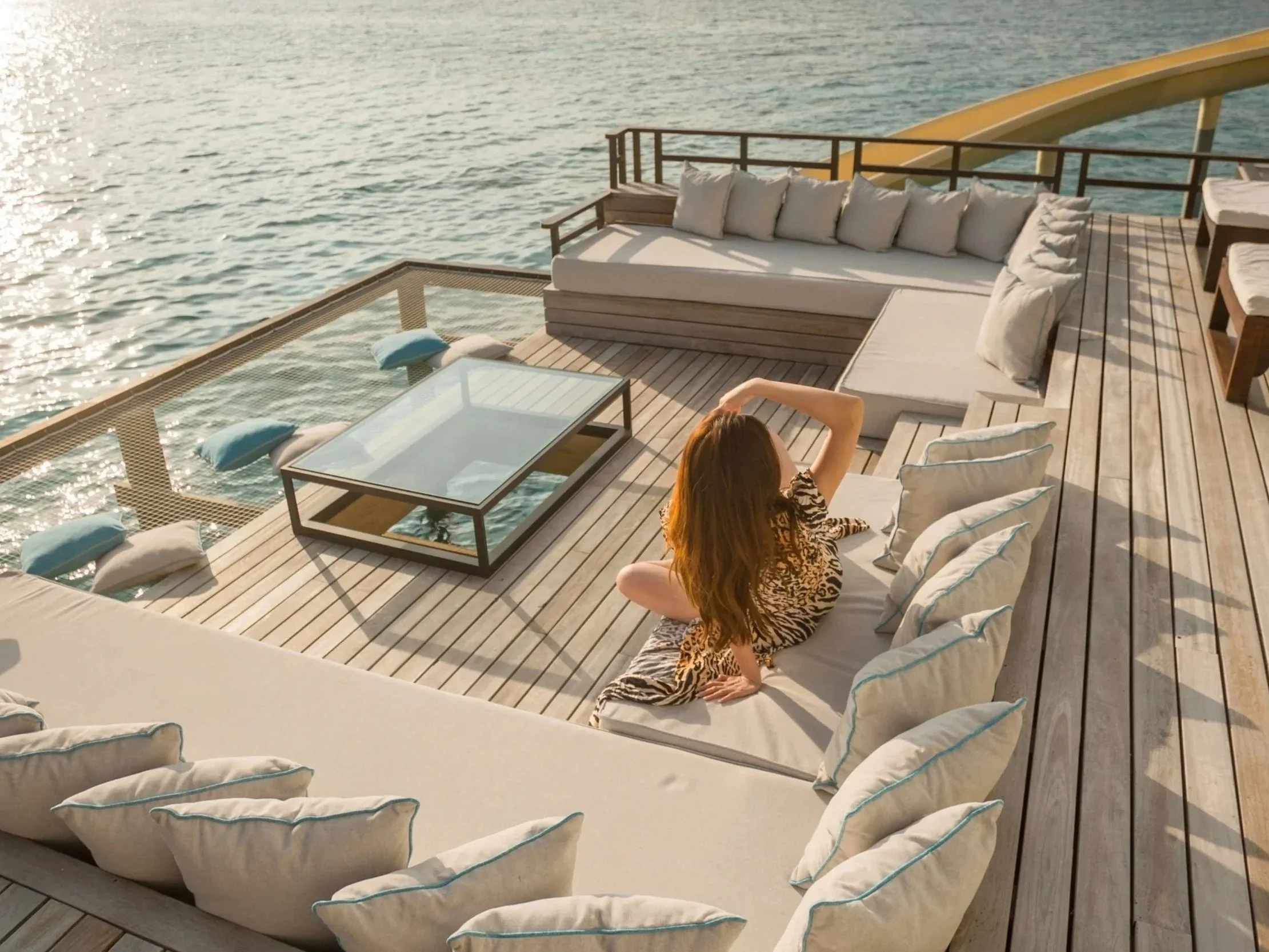 Woman with red hair sitting on a white cushioned lounge chair on a wooden deck by the water, relaxing and enjoying the view.