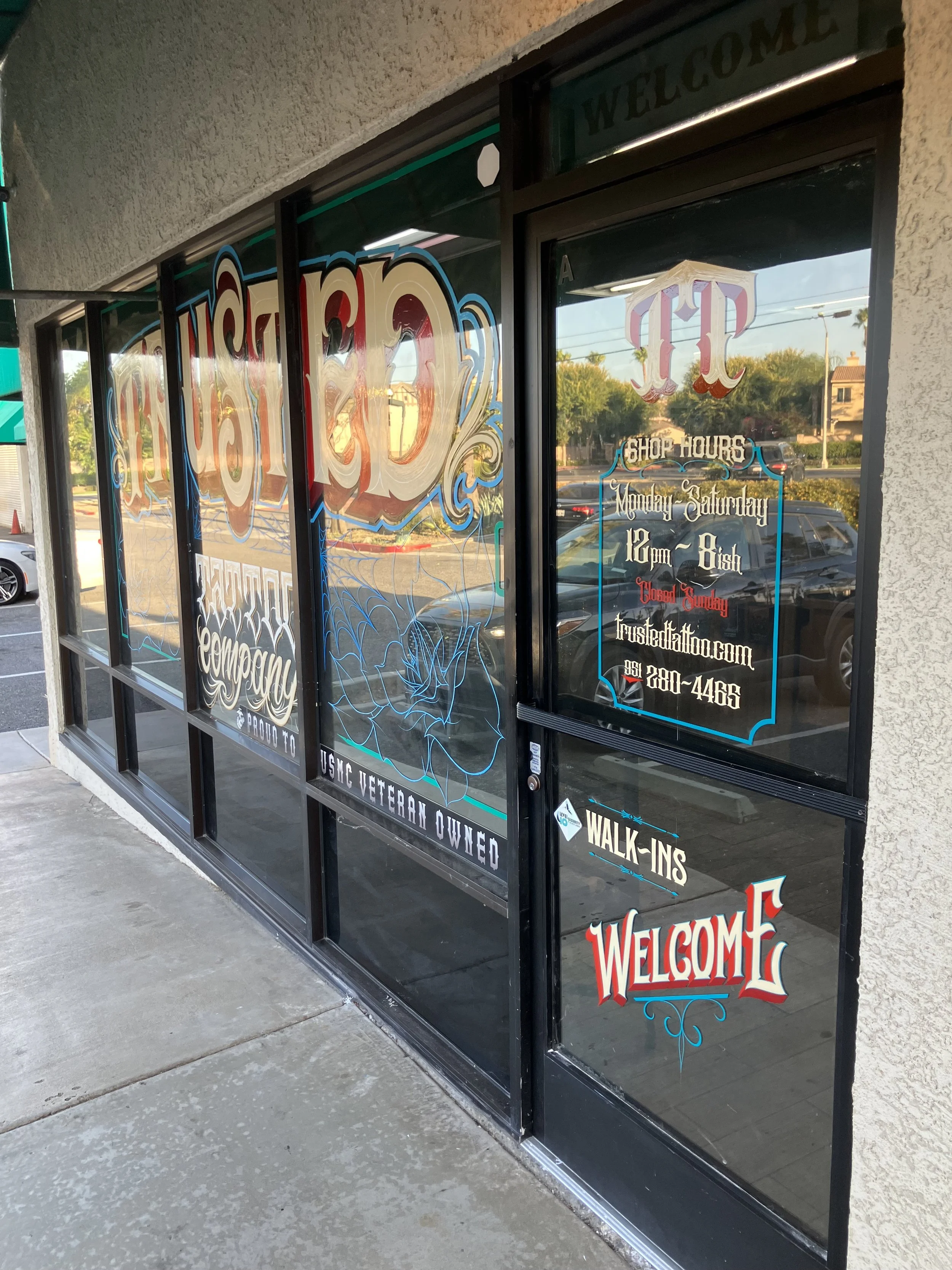 Storefront with welcome signs and decorative lettering, indicating shop hours, contact information, and that the store is closed on Sundays. Displayed on glass doors.