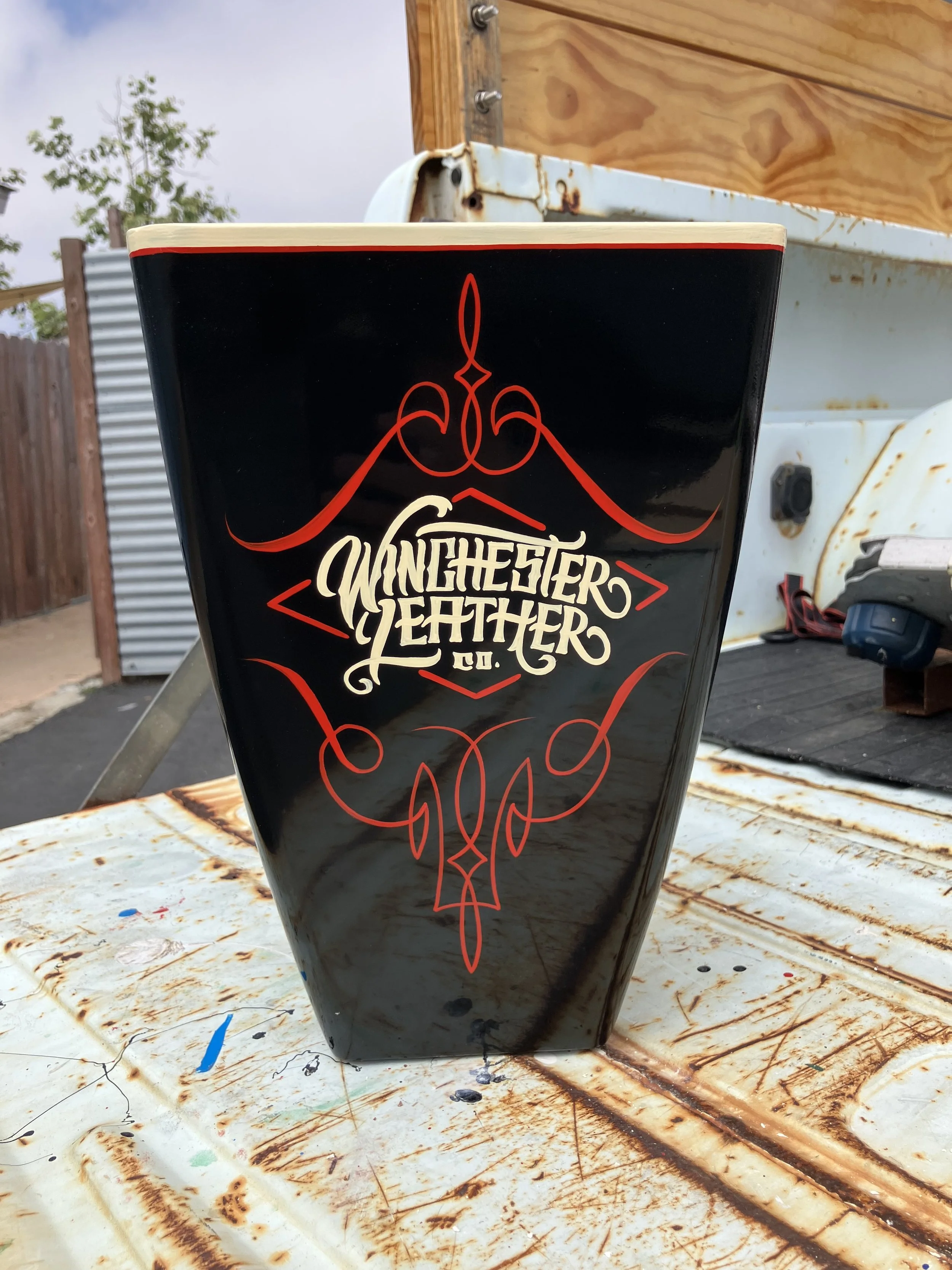 Black mug with a Winchester Leather Co. logo in white lettering and a red decorative design on a rusty surface with wooden background and metal fence.