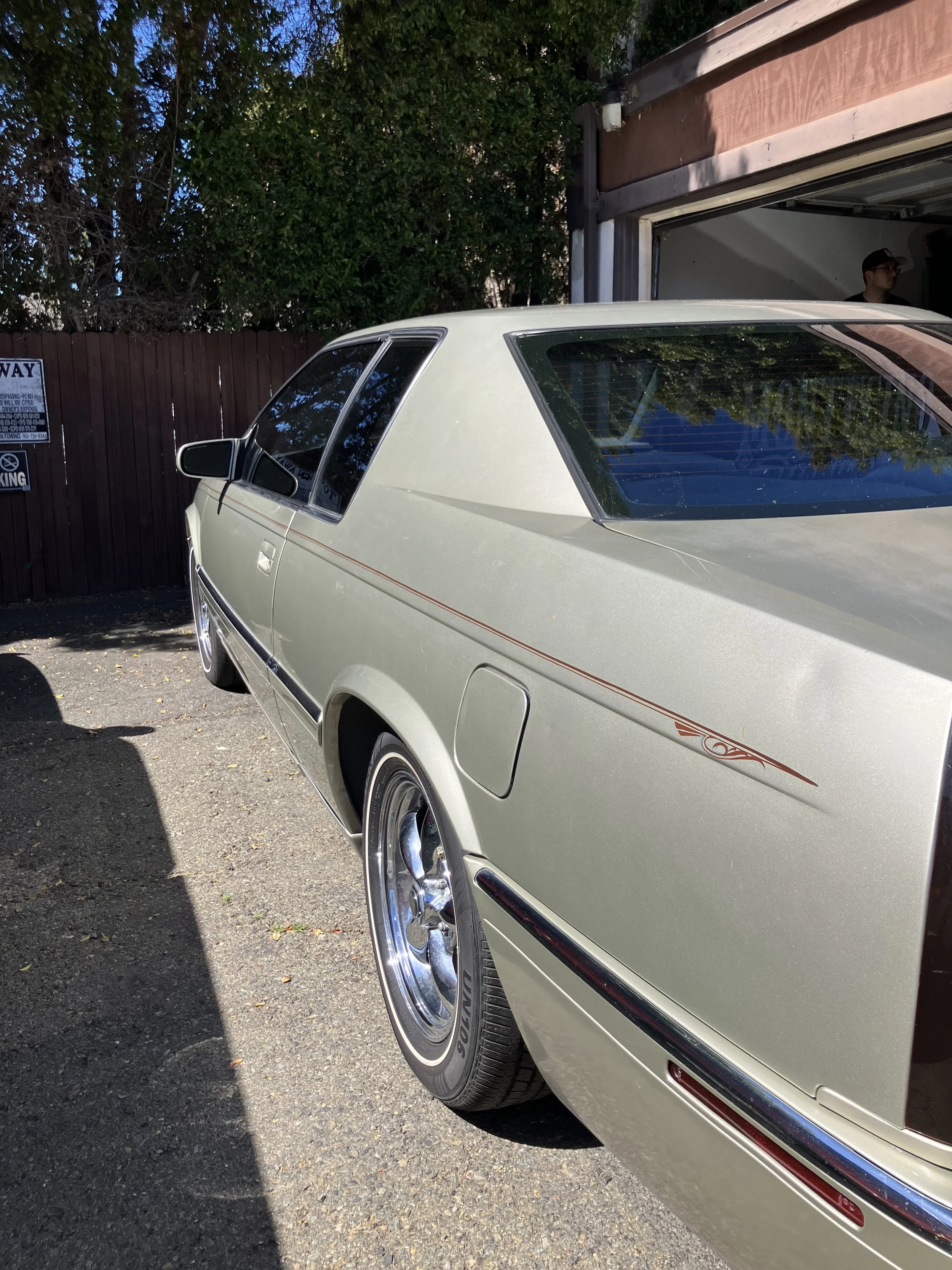 A vintage cream-colored car parked outside a garage with a person standing inside, and trees reflected in the car's rear window.