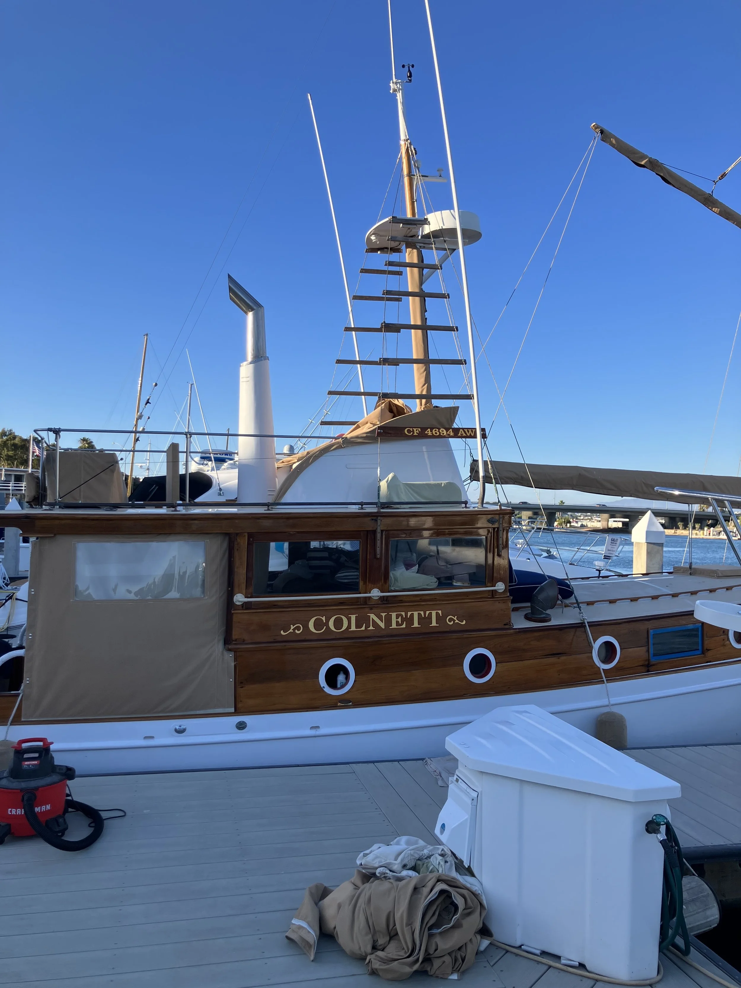 Photo of a boat dock with various items including a laundry pile, a vacuum cleaner, and a white cabinet, against a background of water, blue sky, and other boats.