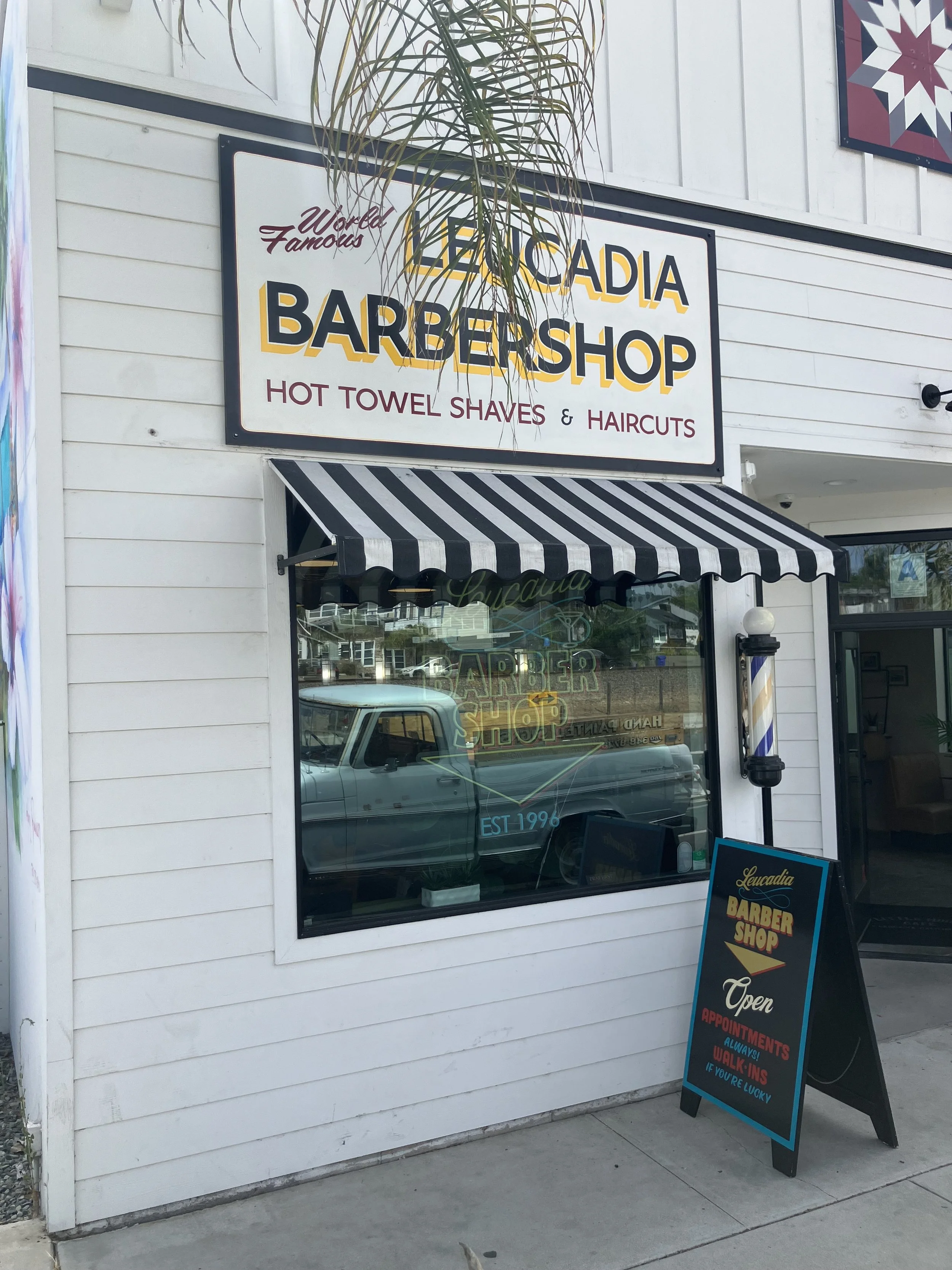 Storefront sign for Loucadia Barber Shop with a black and white striped awning. A sidewalk sign outside indicates the shop is open and accepts appointments, walk-ins always, and if you're lucky. Reflection of a pickup truck in the storefront window.