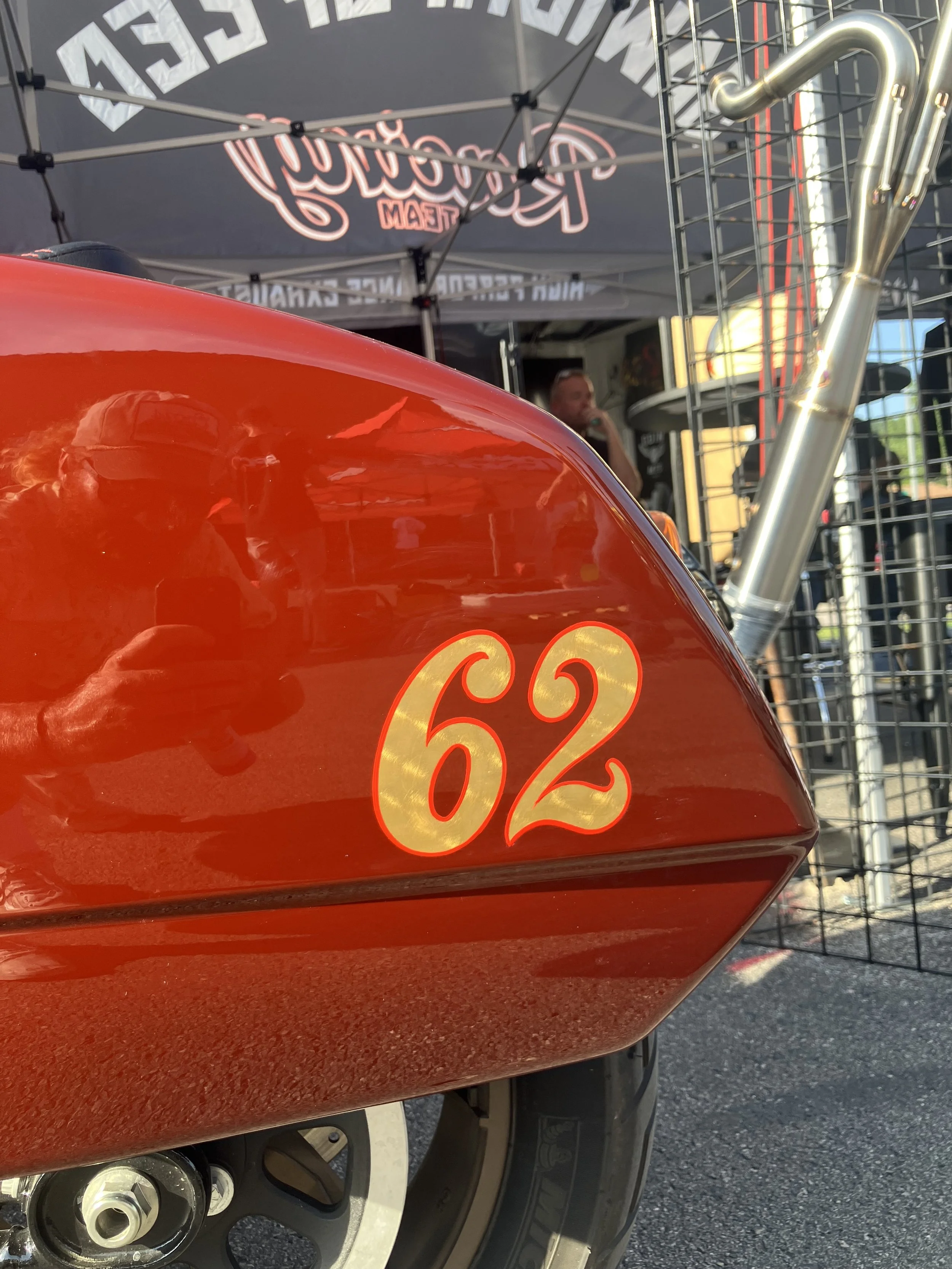 Close-up of a red motorcycle gas tank with the number 62 in gold lettering, part of the front wheel and brake disc visible, a person and a black tent with white and red text in the background.