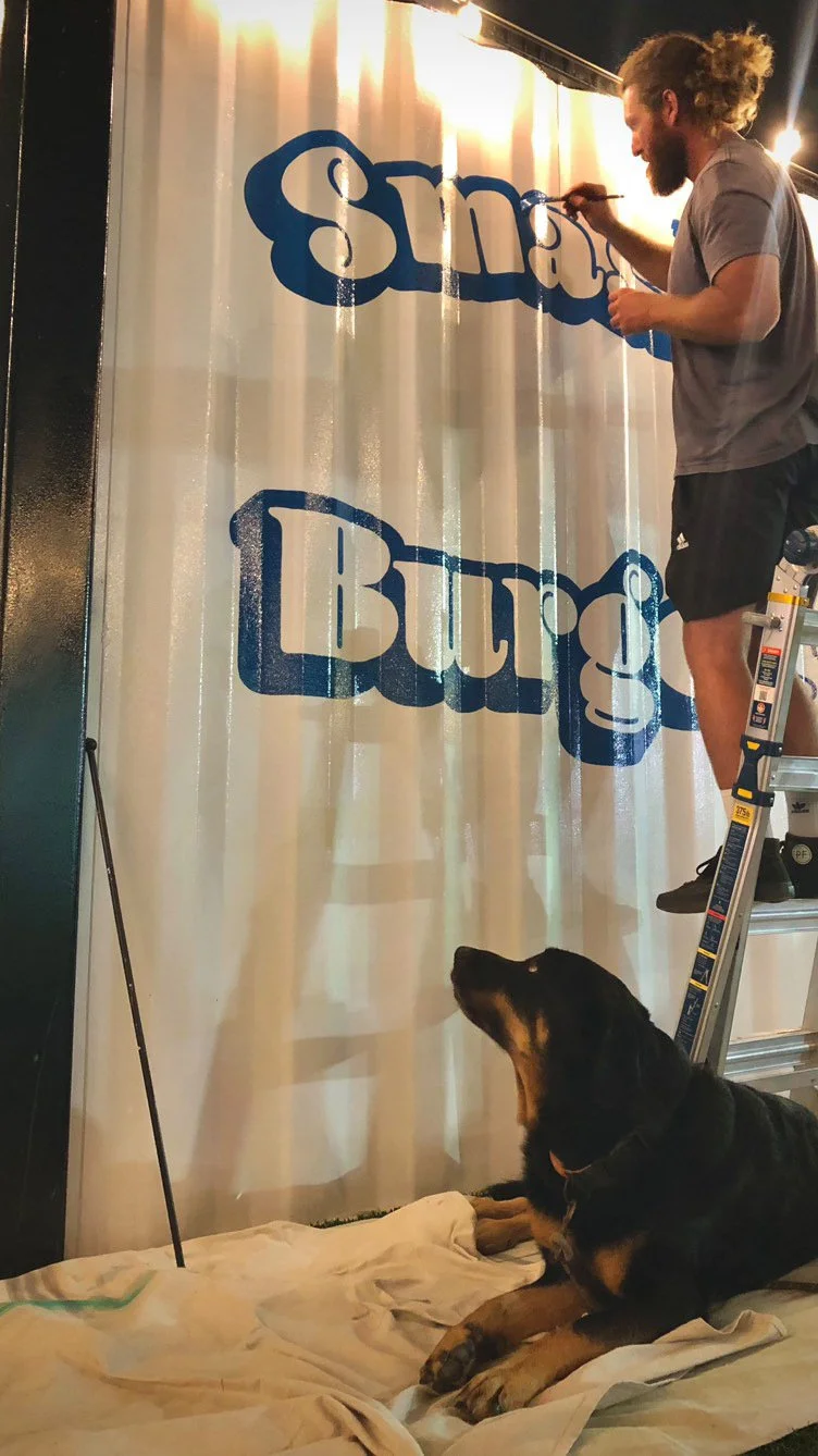 A man on a ladder painting the word "Small" on a wall, while a dog lies on a blanket watching him.