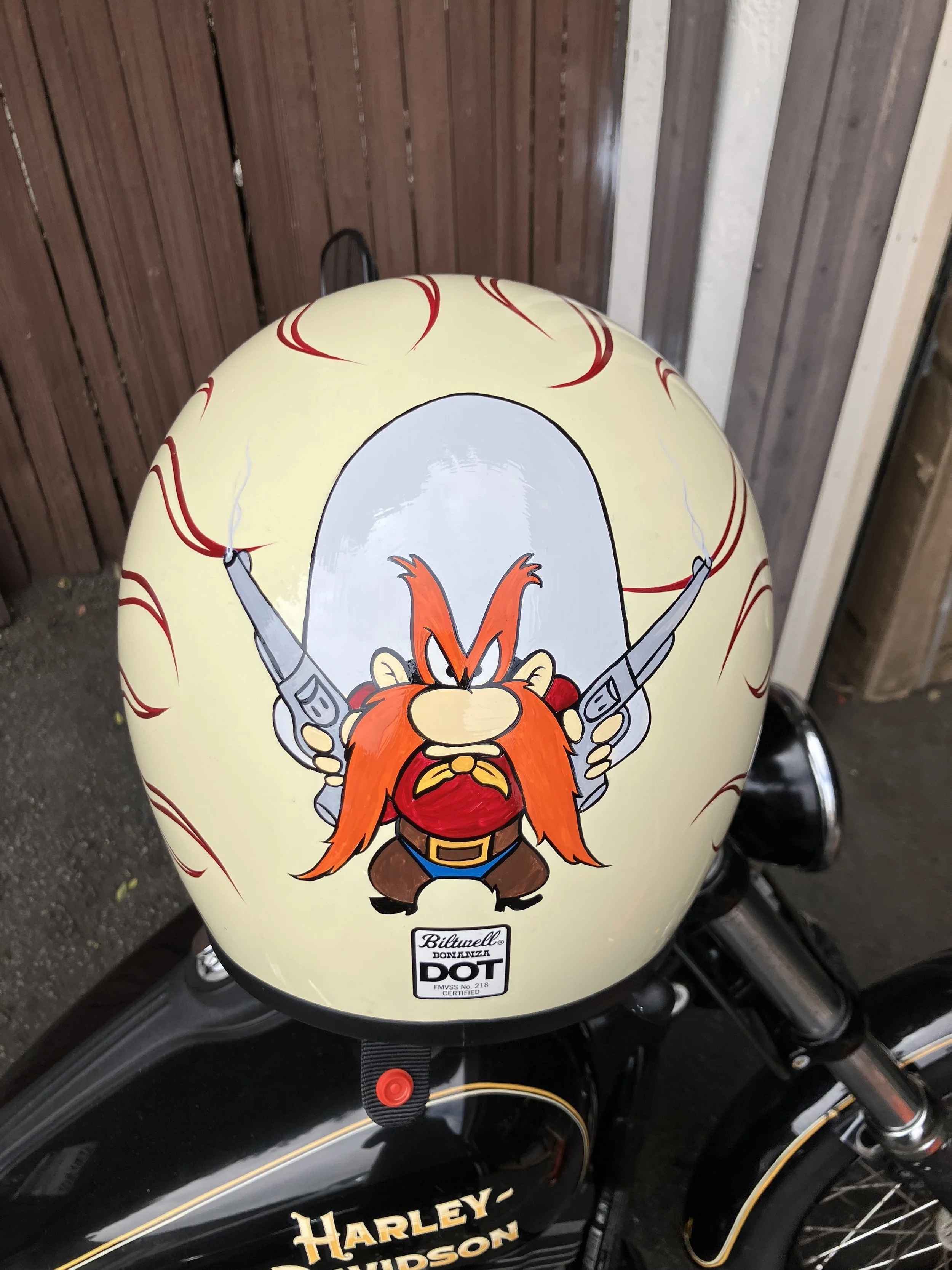 A cream-colored motorcycle fuel tank with a cartoon Viking character holding two thermoses, surrounded by red decorative flames, and the Harley Davidson logo underneath.