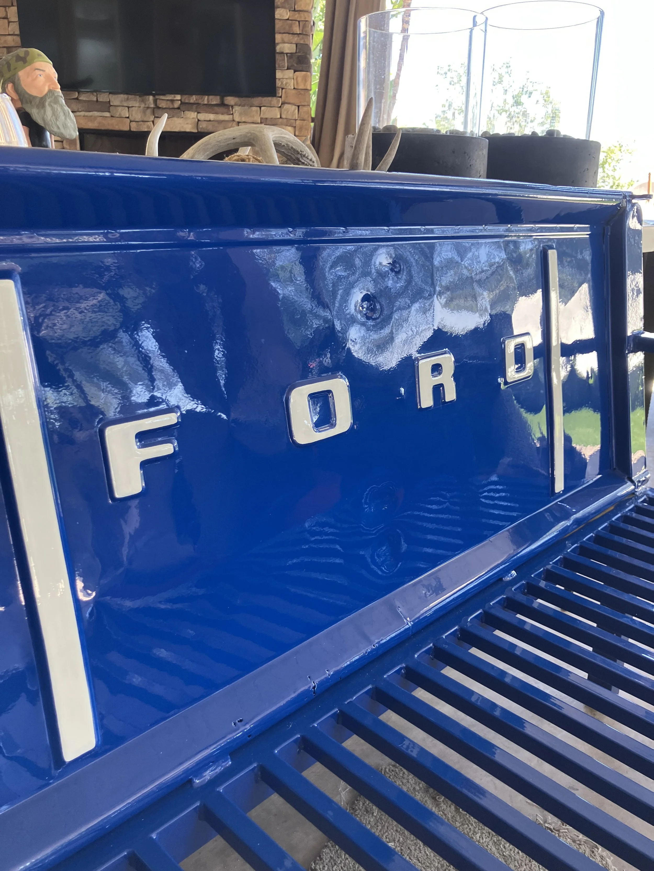 Part of a blue Ford vehicle, possibly a truck or Jeep, with a grille at the bottom and chrome accents spelling out 'FORD'. In the background, there is a decorative display featuring antlers, a bear figurine, and glass candle holders, with a stone fir