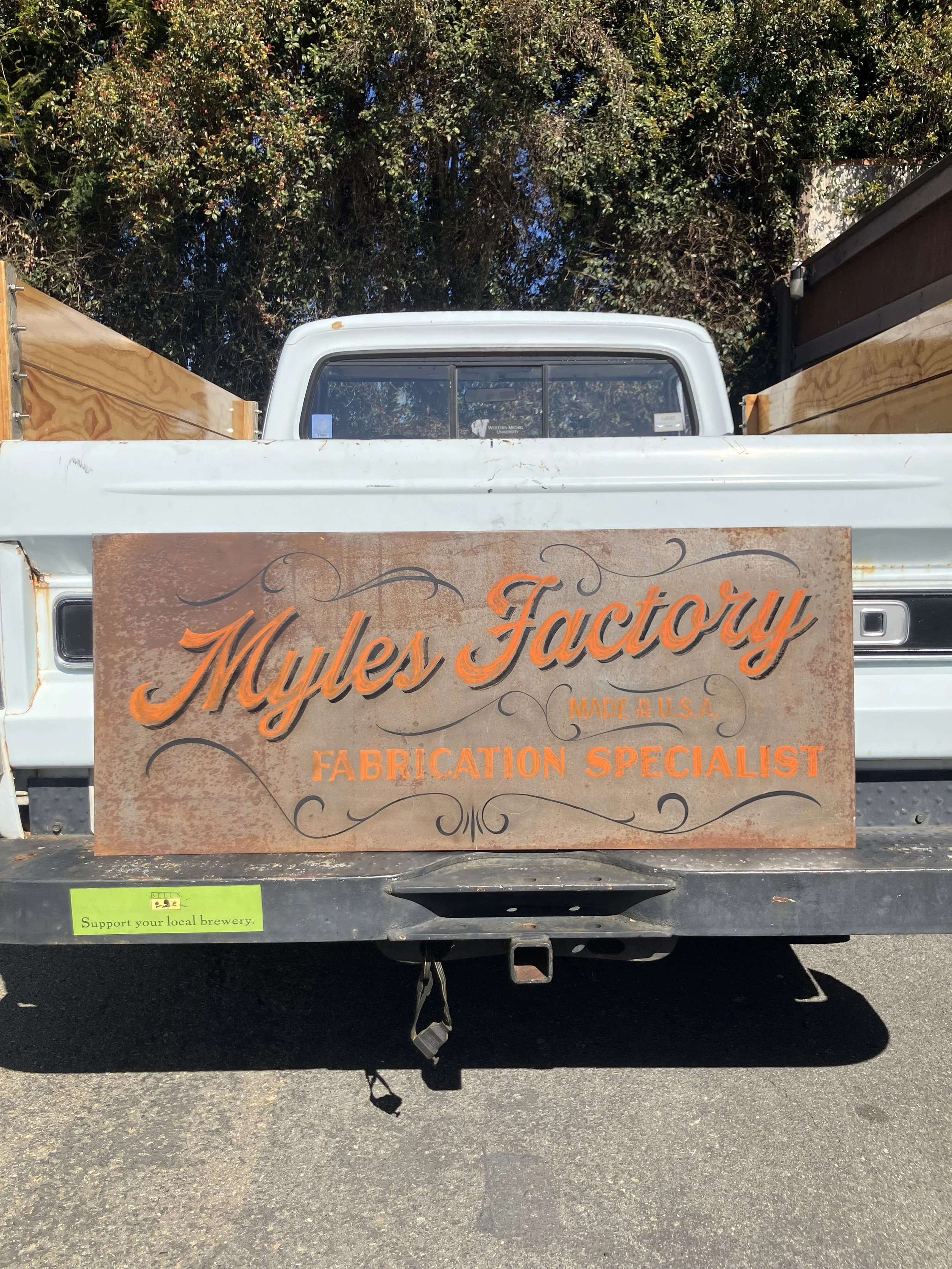 A white pickup truck with a wooden side panel, parked on a street with trees in the background. A sign on the truck reads "Mules Factory" and indicates it is made in the USA, specializing in fabrication. There is also a green sticker that says "Suppo