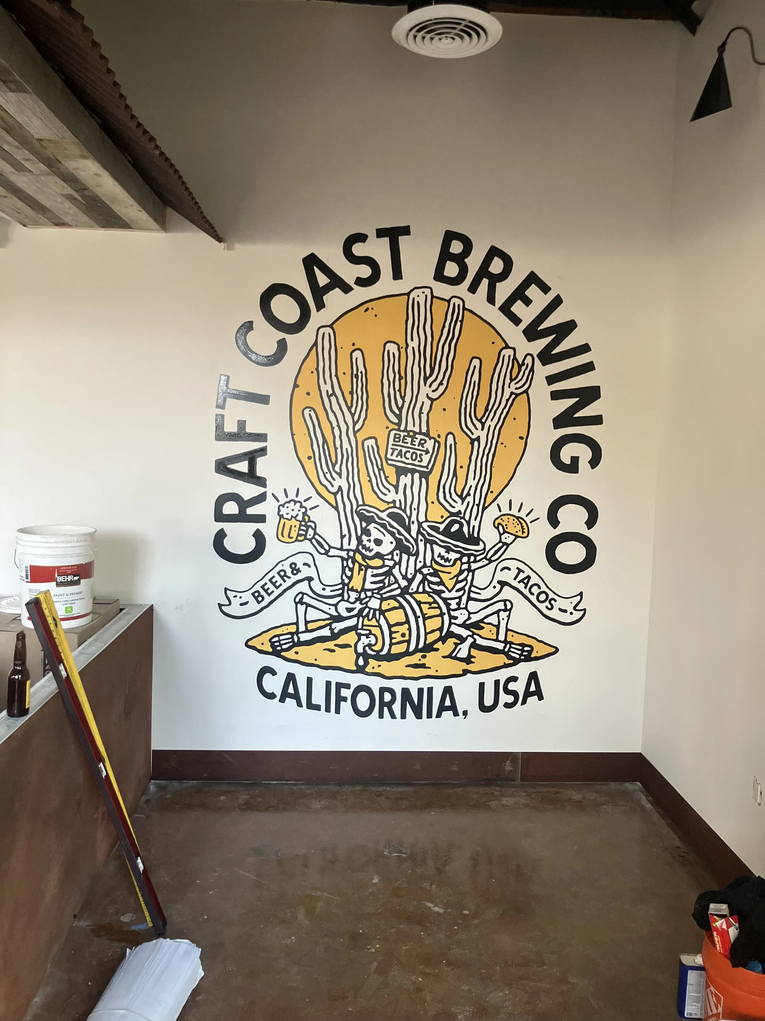 Interior of a restaurant or brewery with a large mural on the wall that says 'CRAFT OAST BREWING CO.' and depicts a cactus holding a beer mug, wearing a hat, and standing on a surfer's surfboard. The mural also says 'BEER & TACOS' and 'CALIFORNIA, US