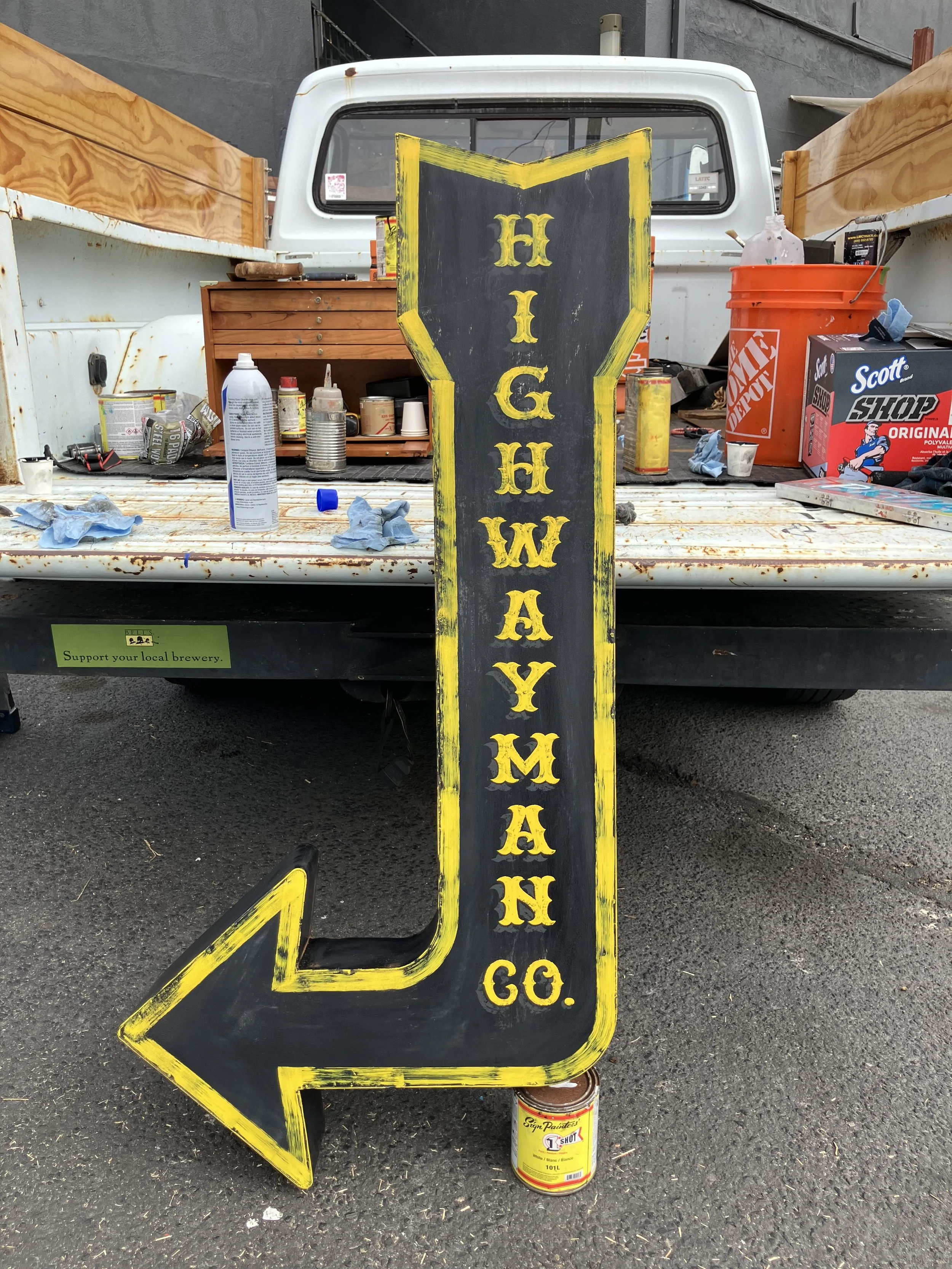A black sign with yellow lettering and border leaning against the back of a white pickup truck. The sign reads 'Highway 66' with a yellow arrow at the bottom pointing left. The truck's bed contains various paint and art supplies, including a can of s