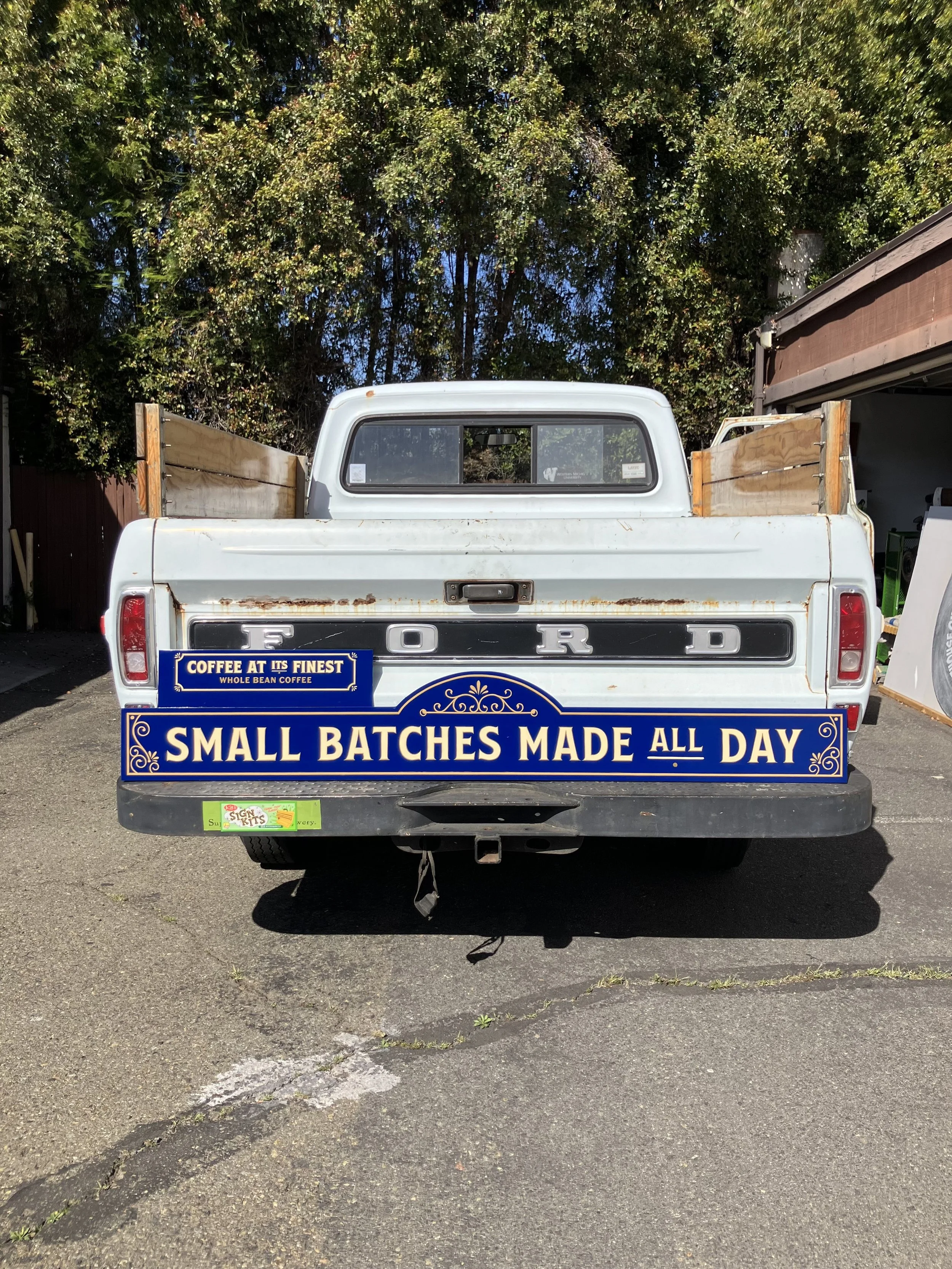 Back of a white Ford pickup truck with a sign that reads, 'Small Batches Made All Day,' parked in a lot with trees behind it and other vehicles nearby.