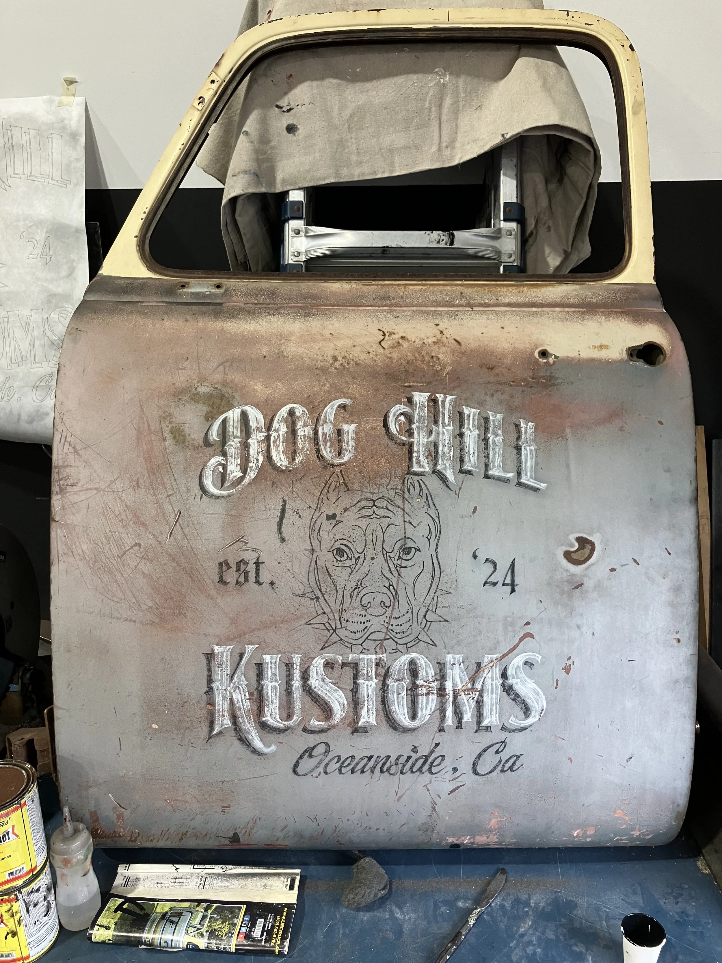 Vintage metal sign reading 'Dog Hill Hustlers Oceanside, Ca' with a dog's face illustration, placed on a workbench surrounded by paint cans, brushes, a magazine, and other art supplies.