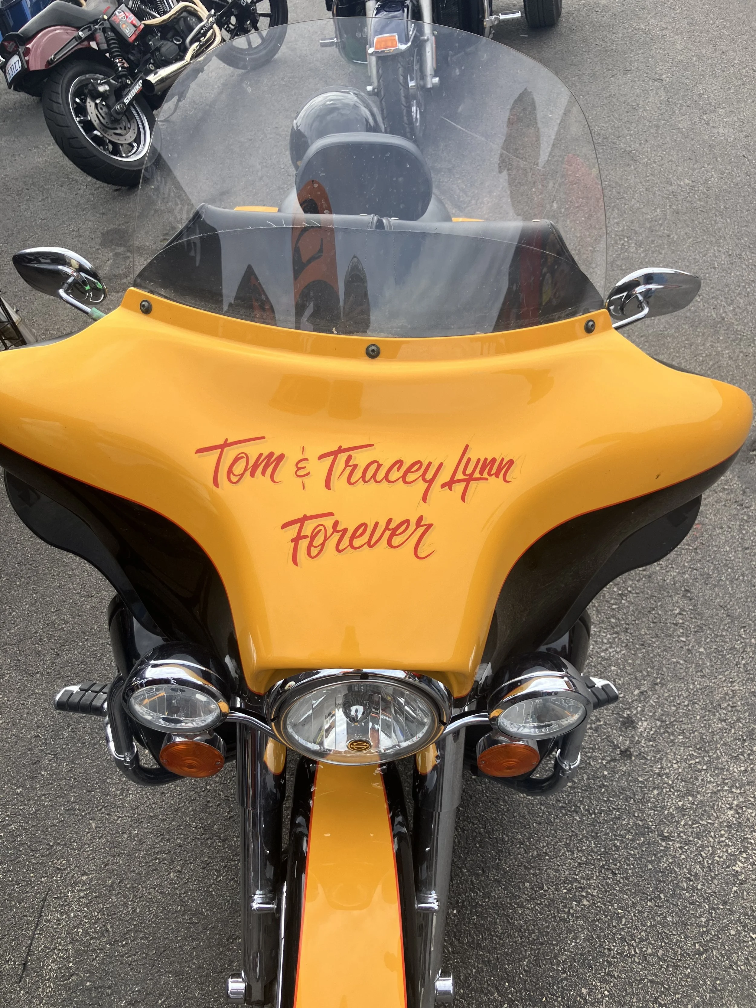 A yellow motorcycle with a windshield and custom writing on the front reading "Tom & Tracey Lynn Forever," parked on a paved surface. There are other motorcycles visible in the background.