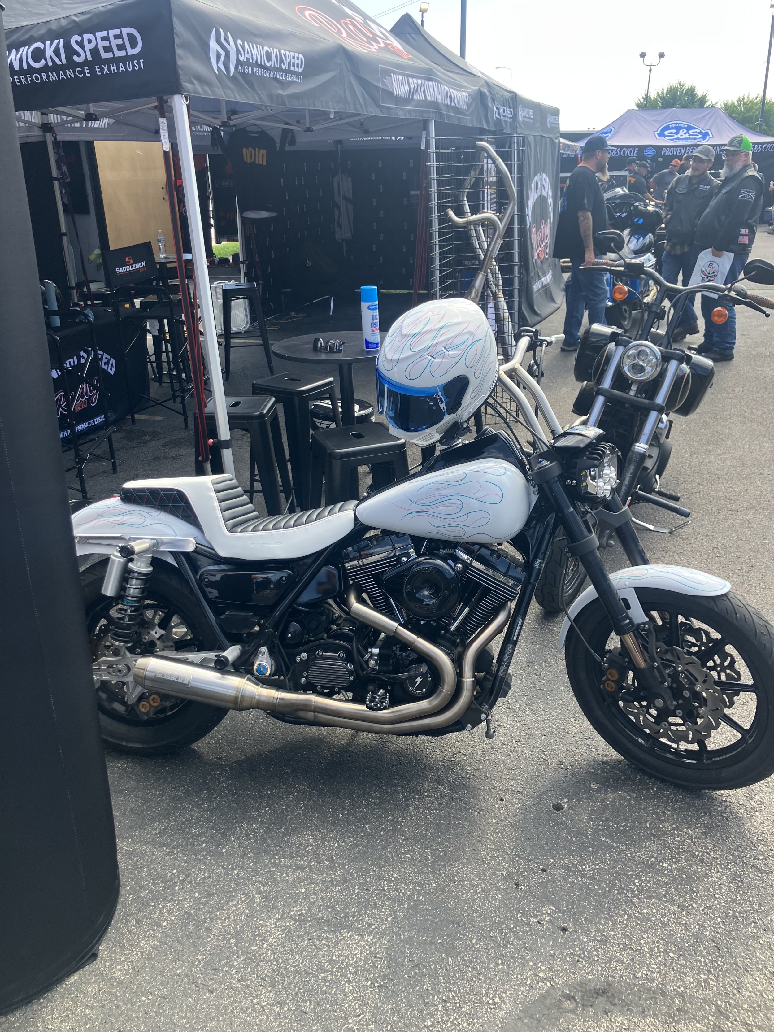 A motorcycle parked in front of event tents at an outdoor venue, with people gathered in the background, and a helmet hanging on the motorcycle handlebar.