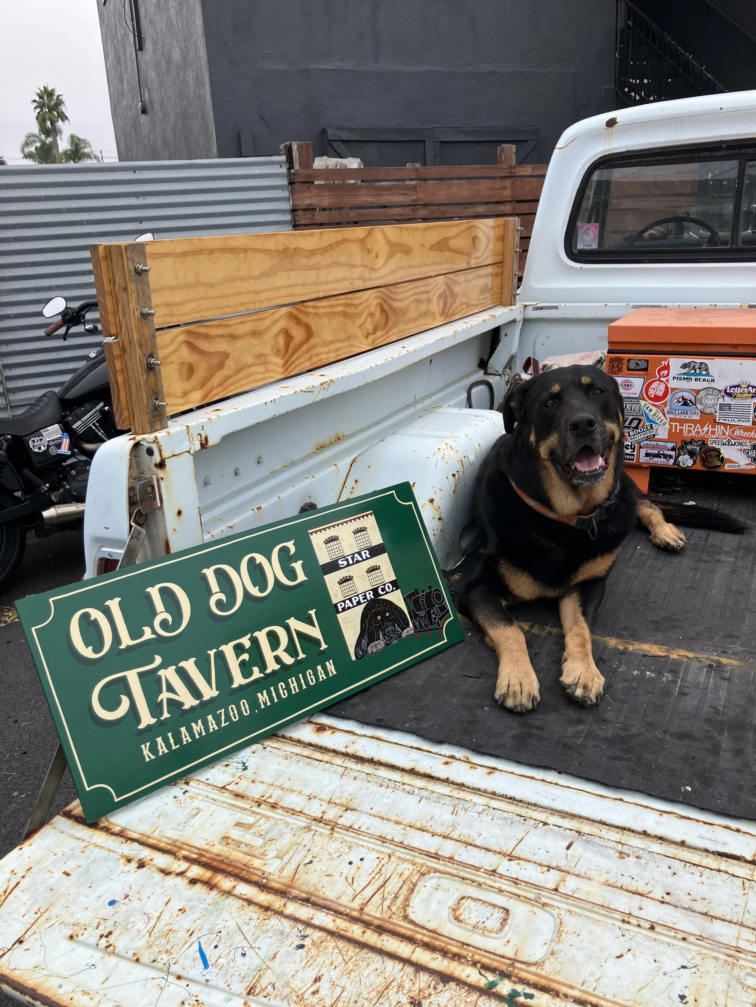 A Rottweiler dog sitting on the back of a rusty white pickup truck with a green and white sign that reads "Old Dog Tavern" leaning against the truck bed. The background includes a black wall, a wooden fence, and some trees.