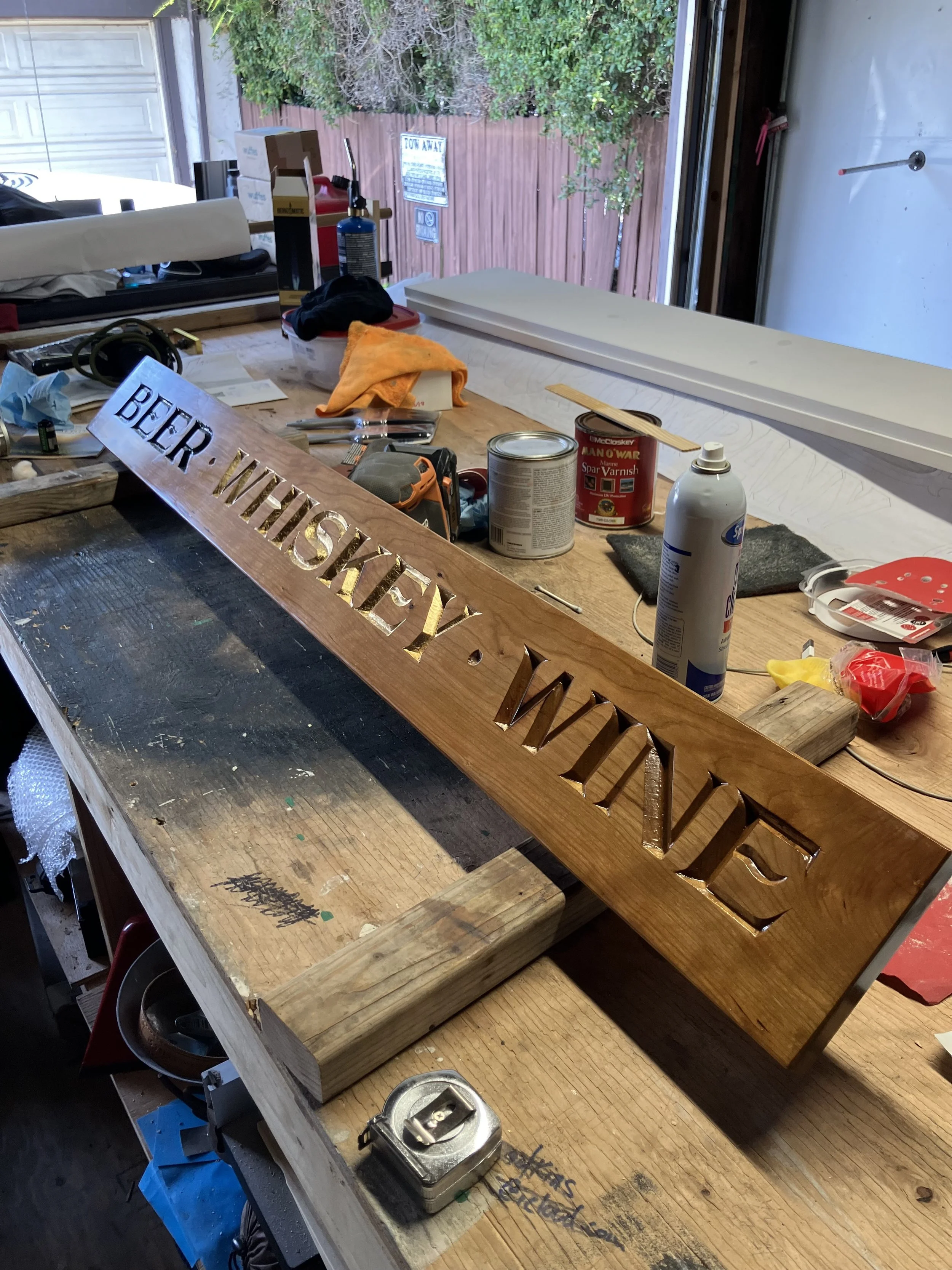 A wooden workbench in a workshop with various tools and supplies, including a metal sign that says 'BEER WISHES' and a piece of wood with cut-out letters. The background shows a garage door and some storage items.