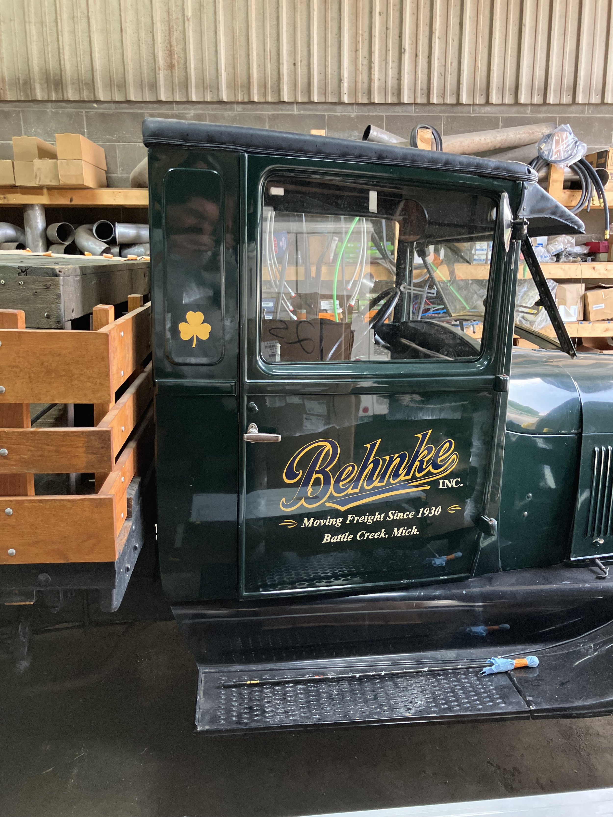A green truck with a wooden flatbed in a warehouse, with a gold shamrock sticker and yellow cursive text on the door that reads "Behnke INC. Moving Freight Since 1930 Battle Creek, Mich." A black rubber mat, a blue work glove, and a screwdriver are on the ground below the truck.