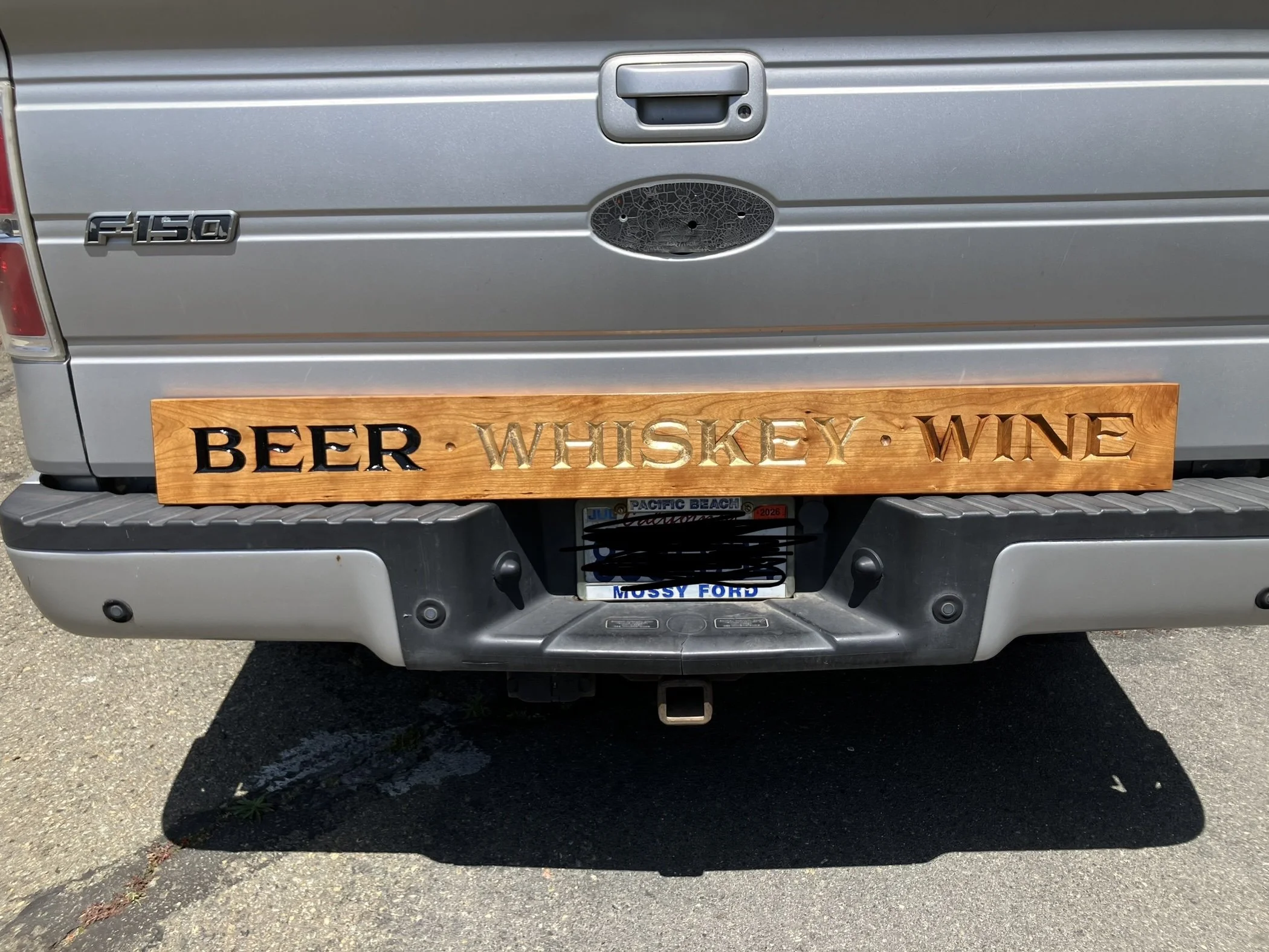 The back of a gray Ford F-150 pickup truck with a wooden sign on the bumper that says 'BEER, WHISKEY, WINE'
