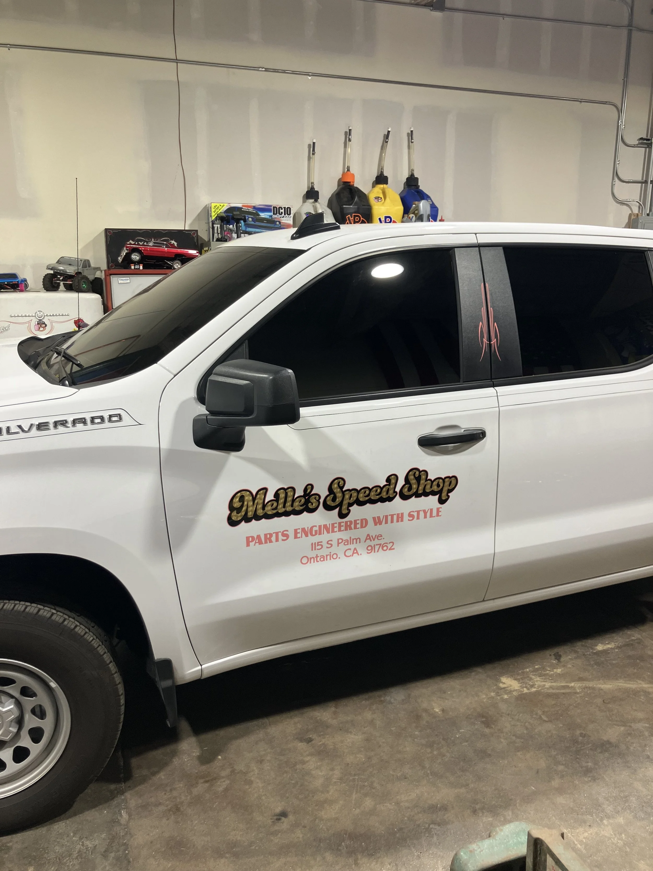 A white Chevrolet Silverado pickup truck with the logo of Melle's Speed Shop on the side, parked indoors. The shop's slogan, 'Parts Engineered with Style,' and address in Ontario, California, are visible on the door. In the background, there are vari