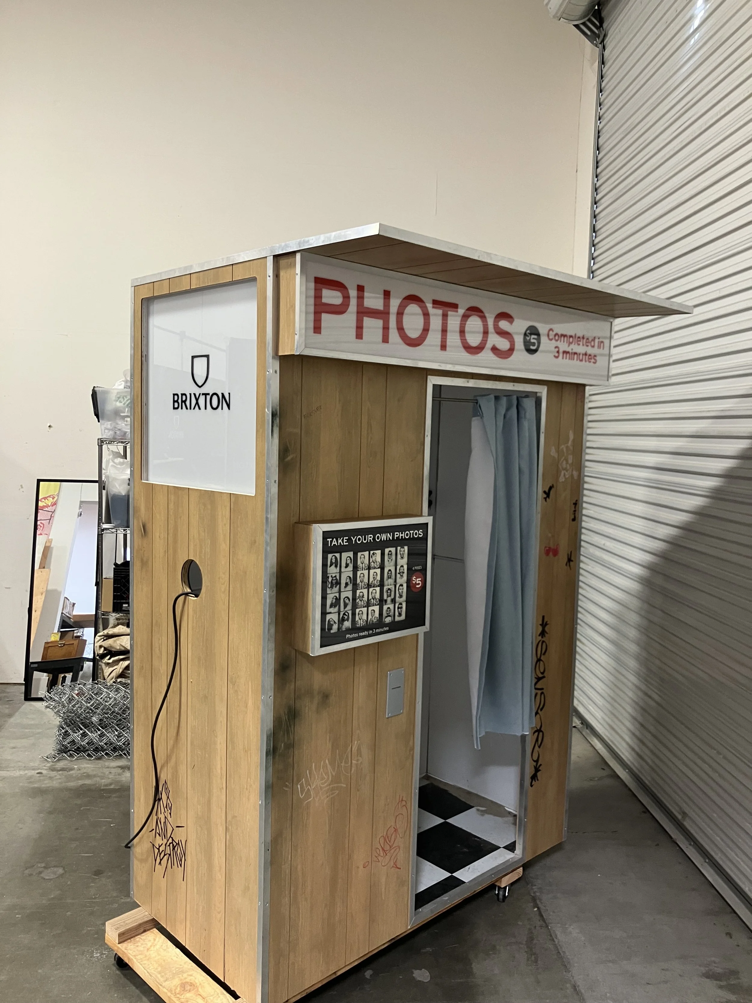 A photo booth with a wooden exterior and a sign that says 'PHOTOS $5 Completed in 3 minutes'. A curtain for photo-taking inside and a framed display with black-and-white portrait photos. The booth is in an indoor space, near a closed metal rolling do