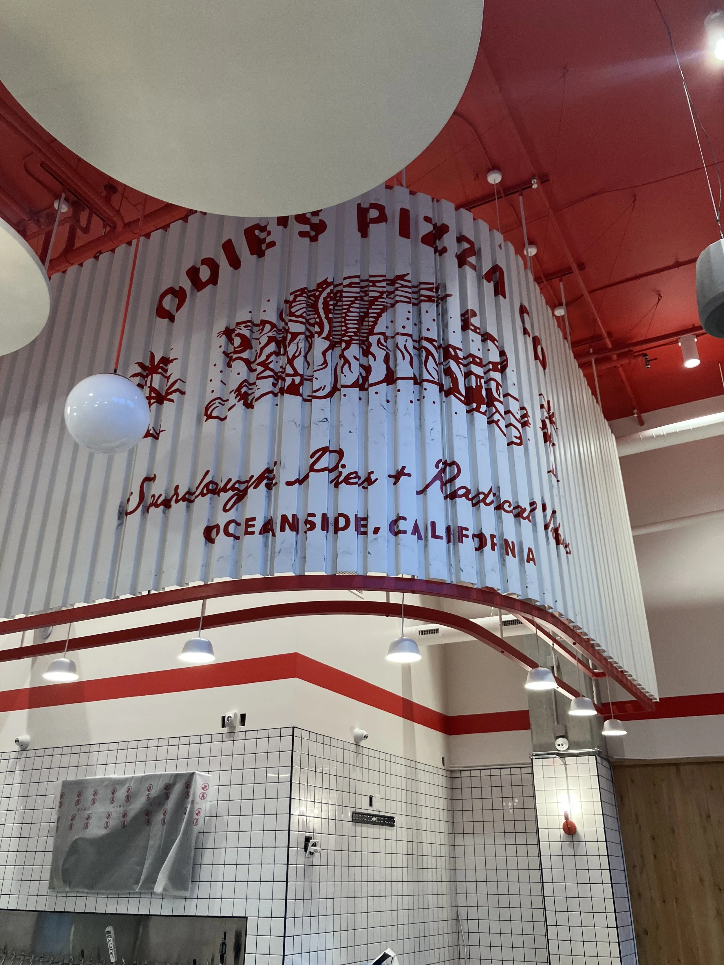 Interior of a modern café or restaurant with white tiled walls, a red curved ceiling, hanging white globe lights, and a decorative white structure with red text and artwork.