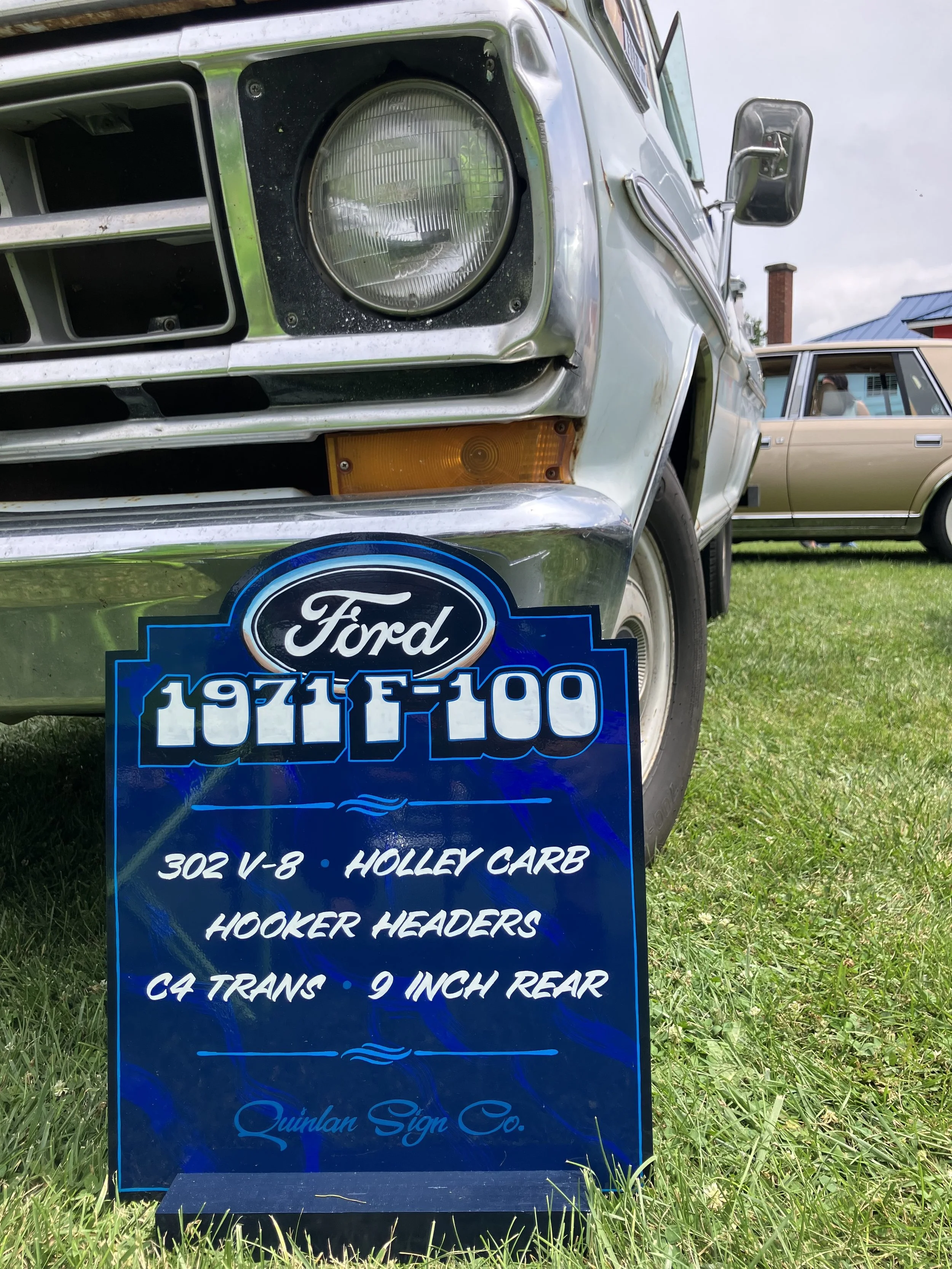 Close-up of a vintage Ford vehicle with a sign in front of it listing specifications like 302 V-8 engine, Holley carburetor, hooker headers, and C4 transmission, displayed outdoors on grass with another similar vehicle in the background.