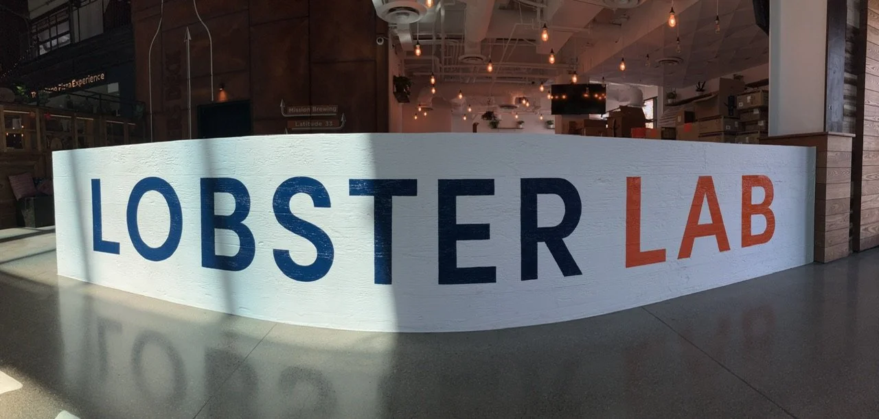 Indoor sign reading 'Lobster Lab' with blue and orange letters, inside a modern space with hanging lights and wooden accents.