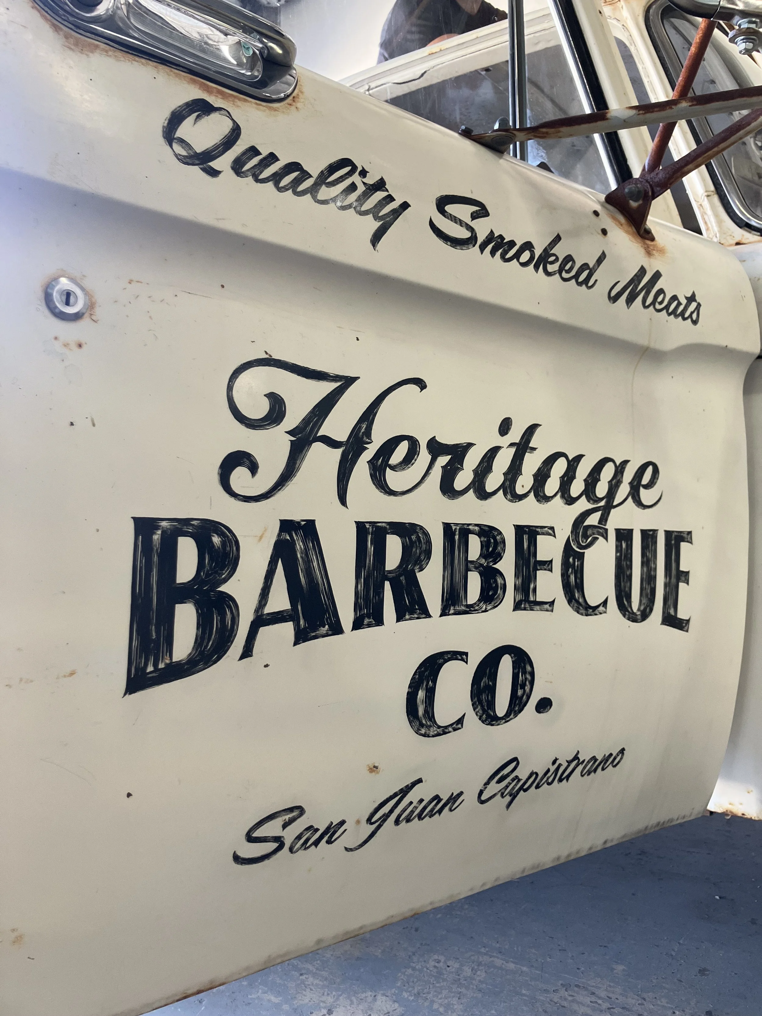 Close-up of a vintage vehicle sign with black handwritten text on a white background that reads: "Quality Smoked Meats" and "Barbecue". The sign appears to be old with some rust spots and scratches.