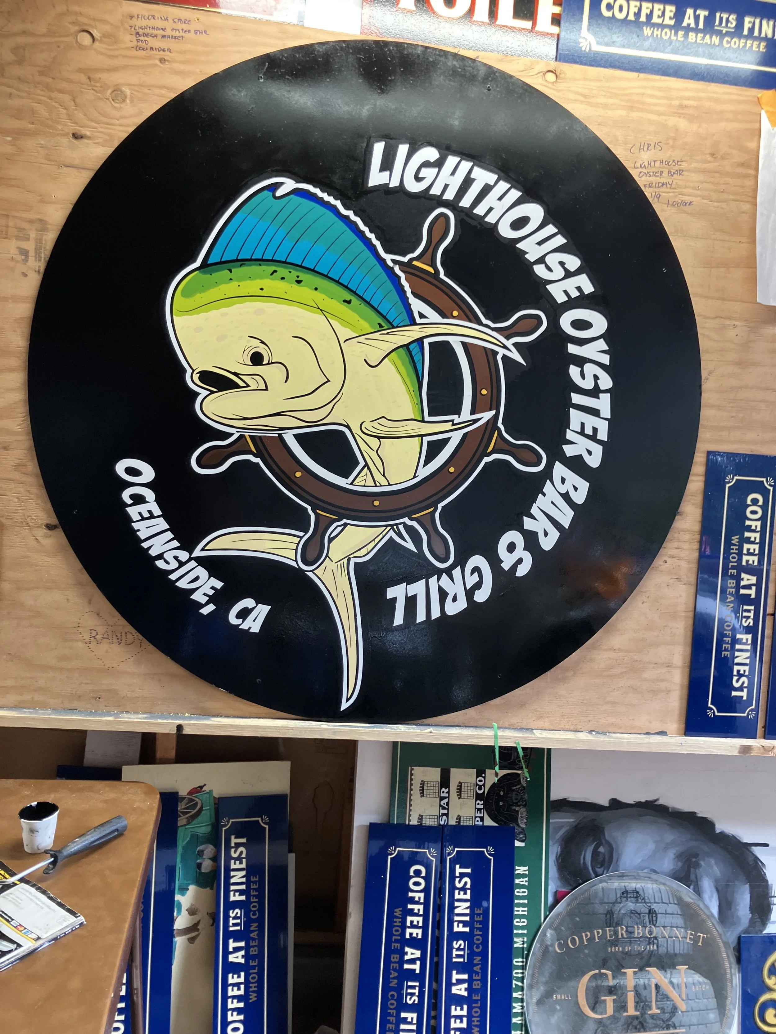 Decorative round sign with a fish illustration and the text 'Lighthouse of Big Sur, CA'. Surrounding the sign are various signs and items, including boxes labeled 'Finest Coffee' and a wooden table with a cup, a marker, and a souvenir plate.