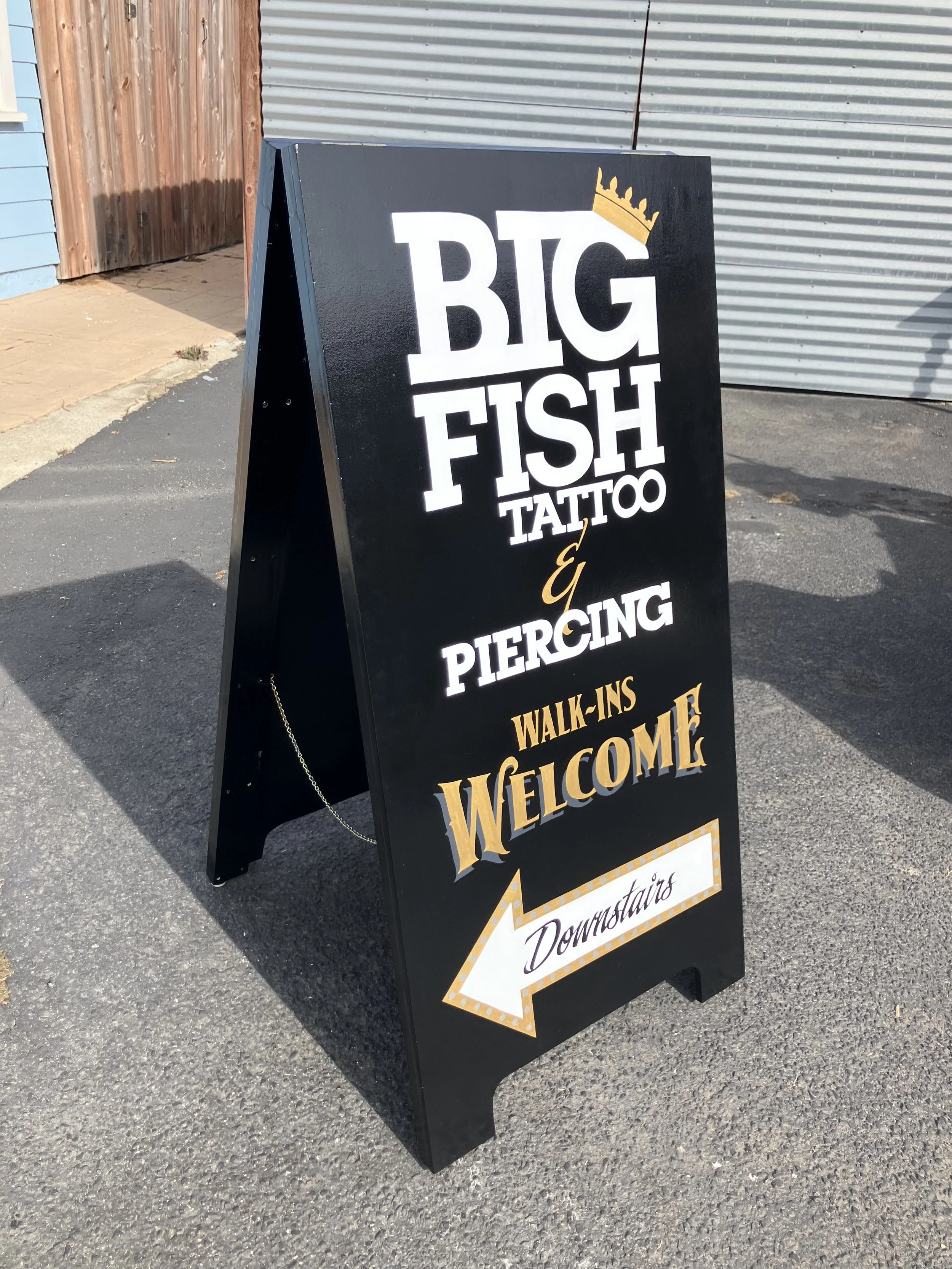 A black standing sidewalk sign advertising a business called Big Fish Tattoo & Piercing with a yellow crown graphic on top of the text, and a yellow arrow at the bottom pointing left with the word Downtown in purple cursive.