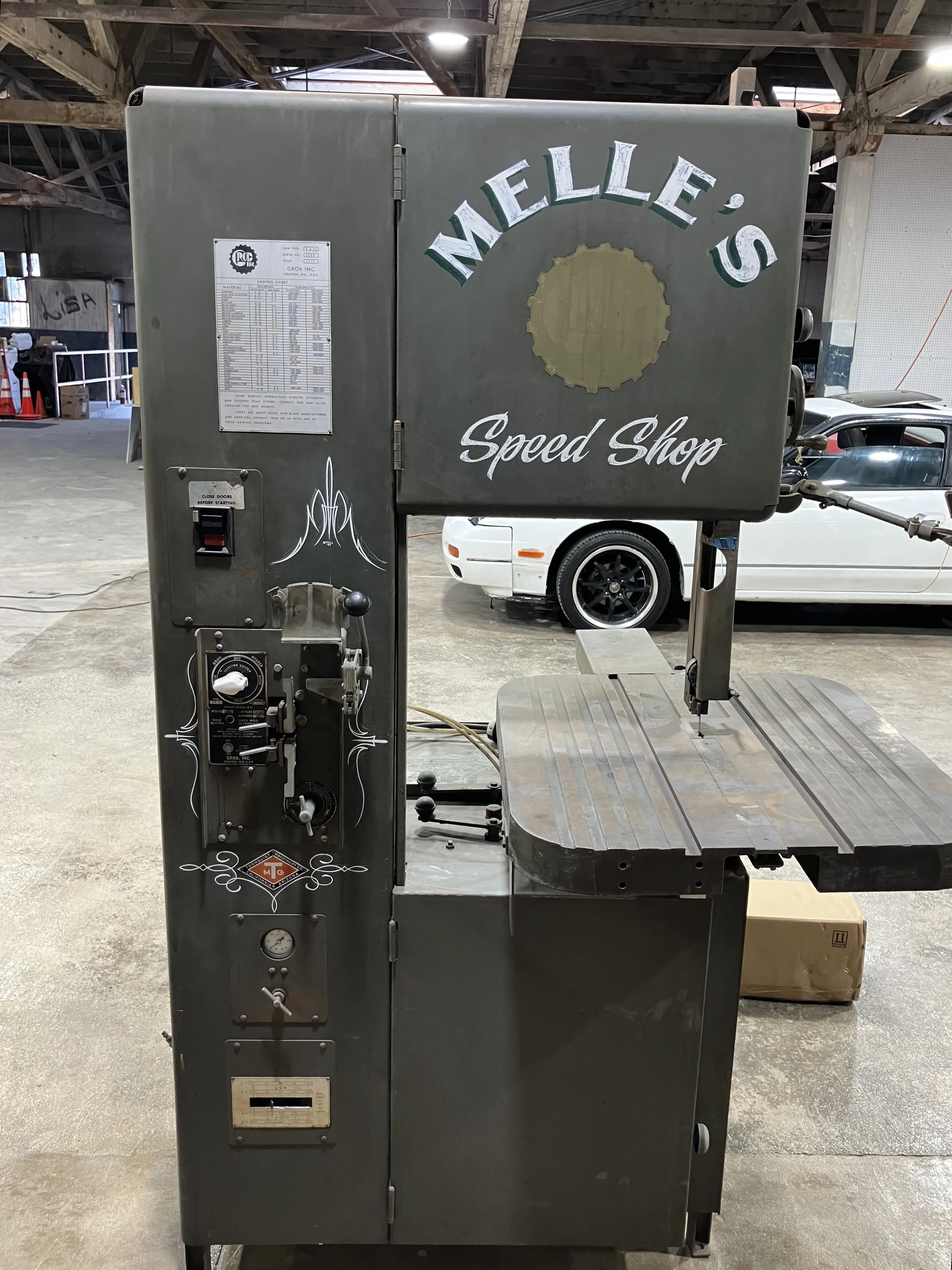 A metal speed shop machine with a sign that reads 'Nalle's Speed Shop'. It has various controls on the side and a large flat working surface. The background shows a warehouse or garage with cars and construction cones.