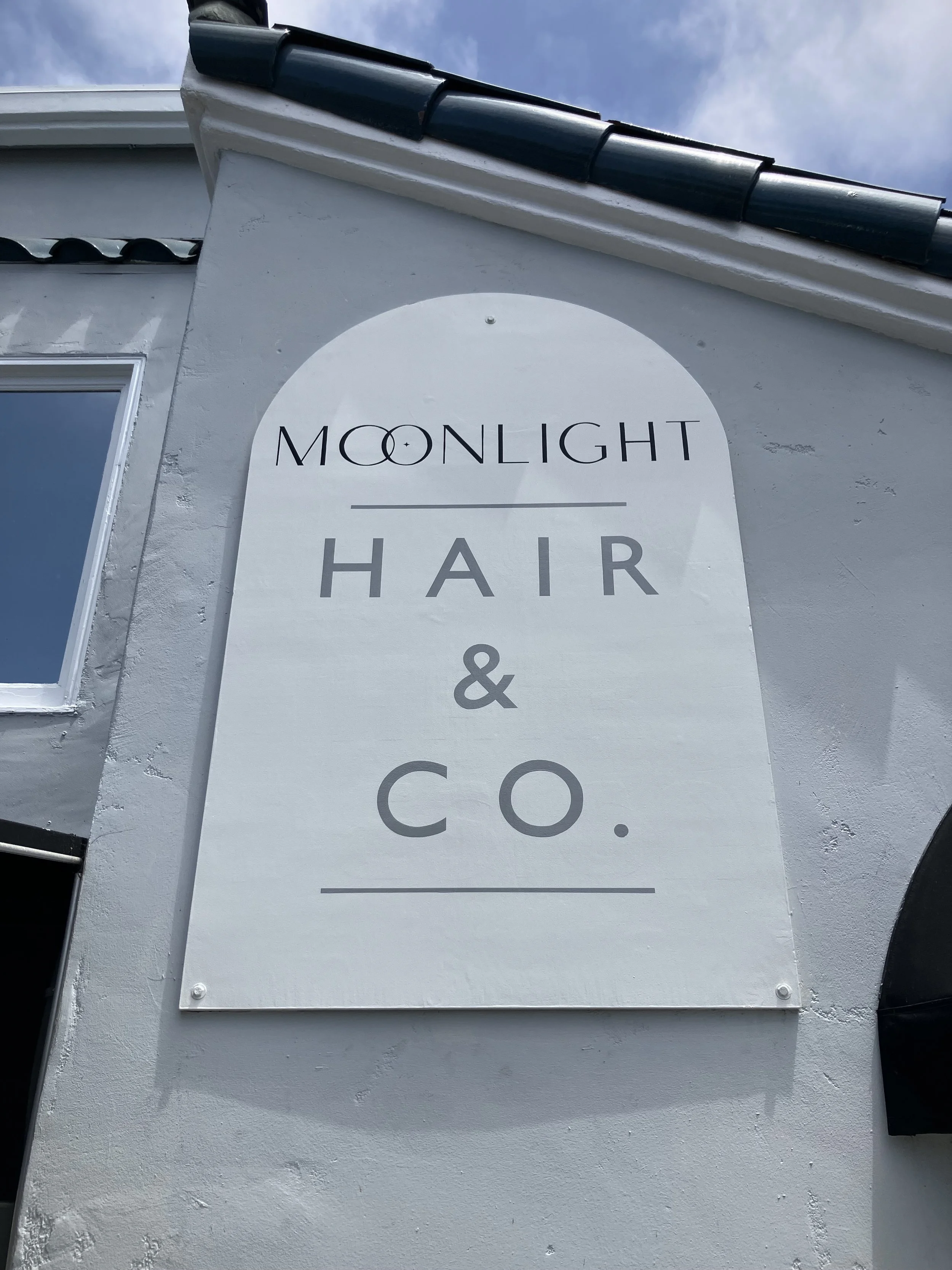 Exterior sign for a business named Moonlight Hair Co. on a white wall, with a window visible on the left and a roof with tiles at the top.