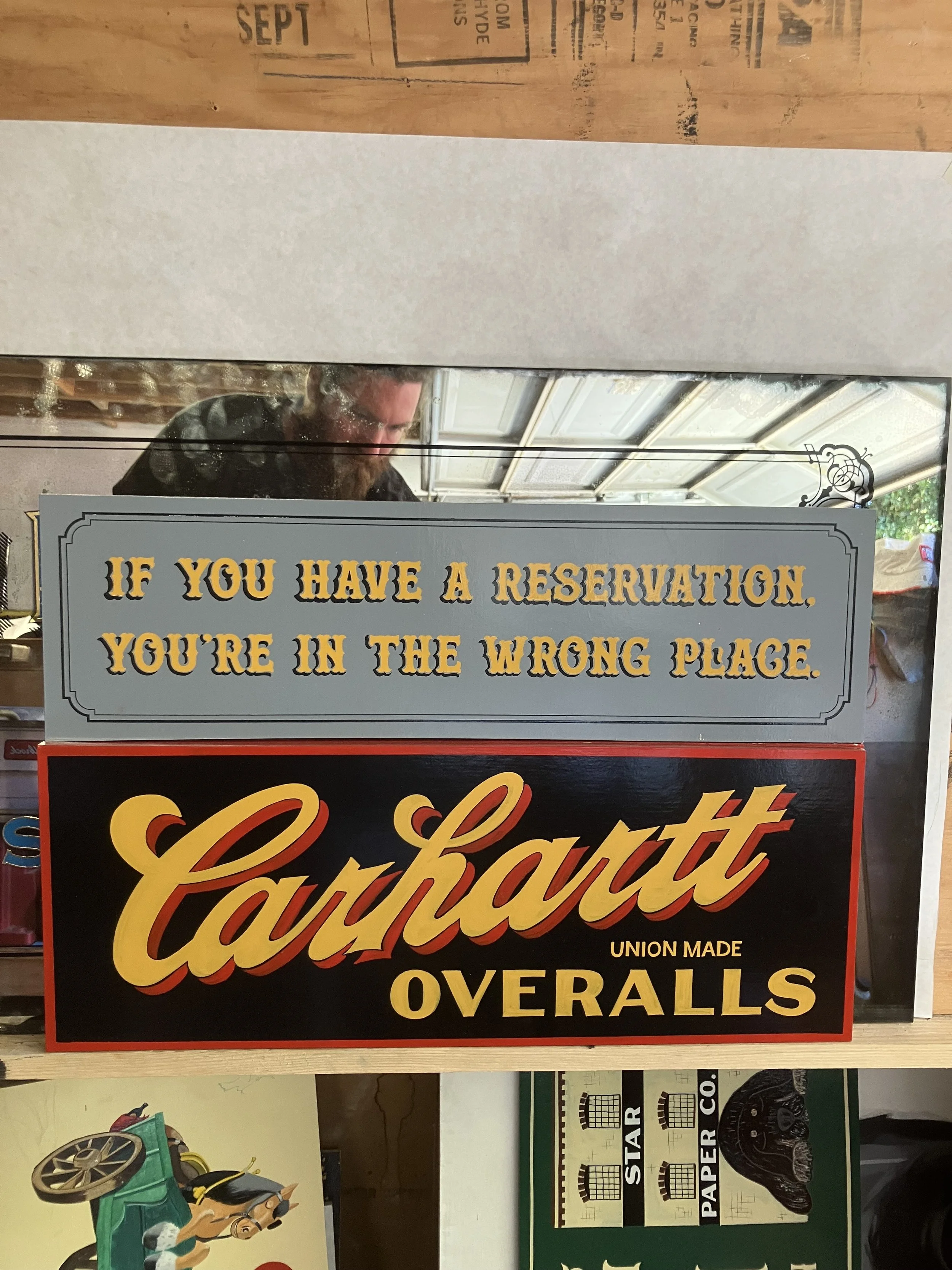 The image shows a sign with a humorous message in yellow and black text on a light gray background, warning that if you have a reservation you are in the wrong place. Behind the sign, parts of a cluttered garage or workshop are visible, including a p