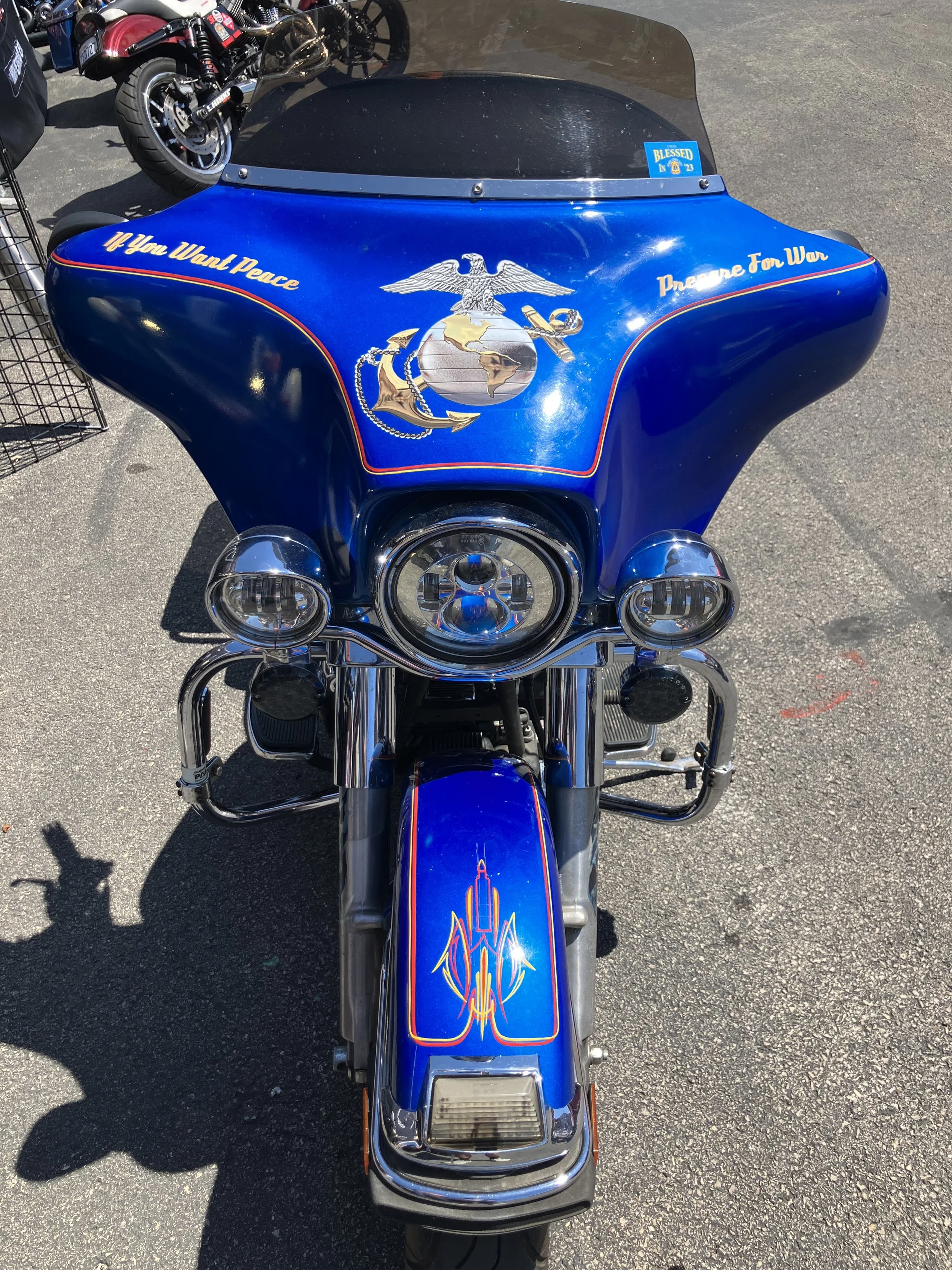 Front view of a blue motorcycle with peace and war-themed stickers, including an eagle, globe, and anchor emblem, and slogan decorations.